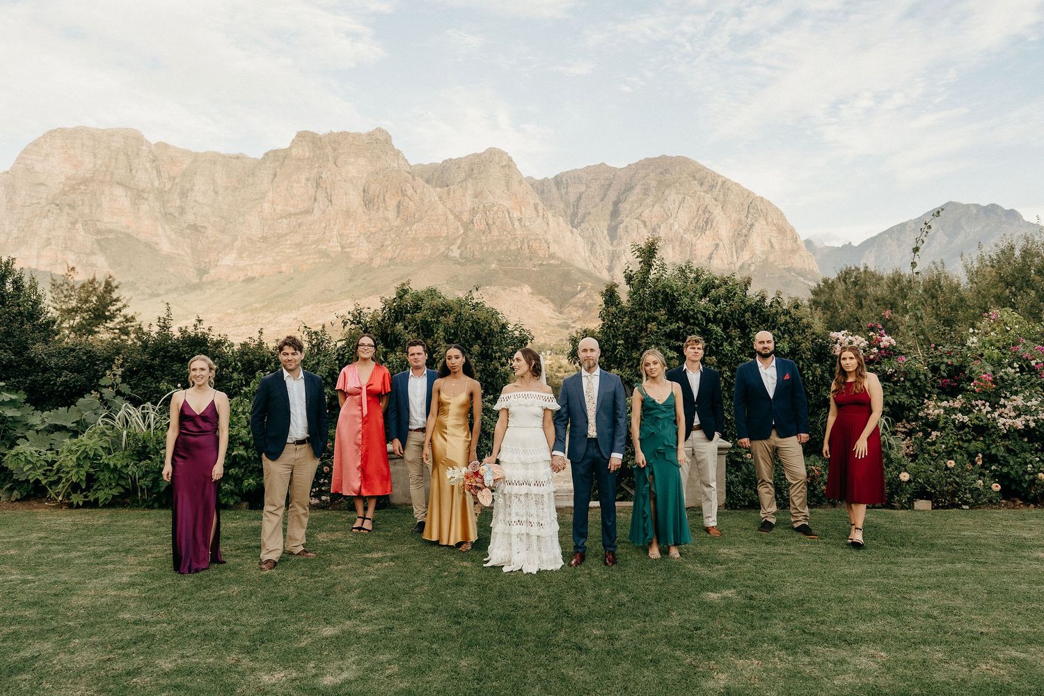 Wedding party poses on green lawn with dramatic mountain peaks and desert landscape as backdrop in scenic outdoor setting.