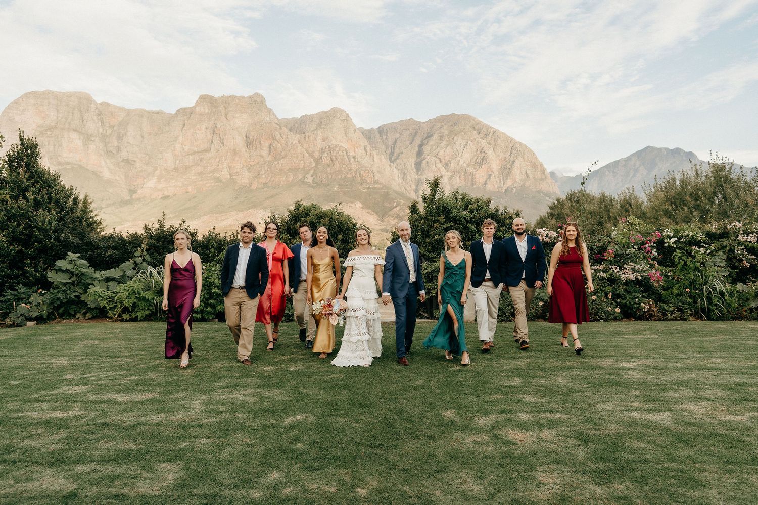 A wedding party stands on a manicured lawn with dramatic mountain peaks rising in the background on a cloudy day.