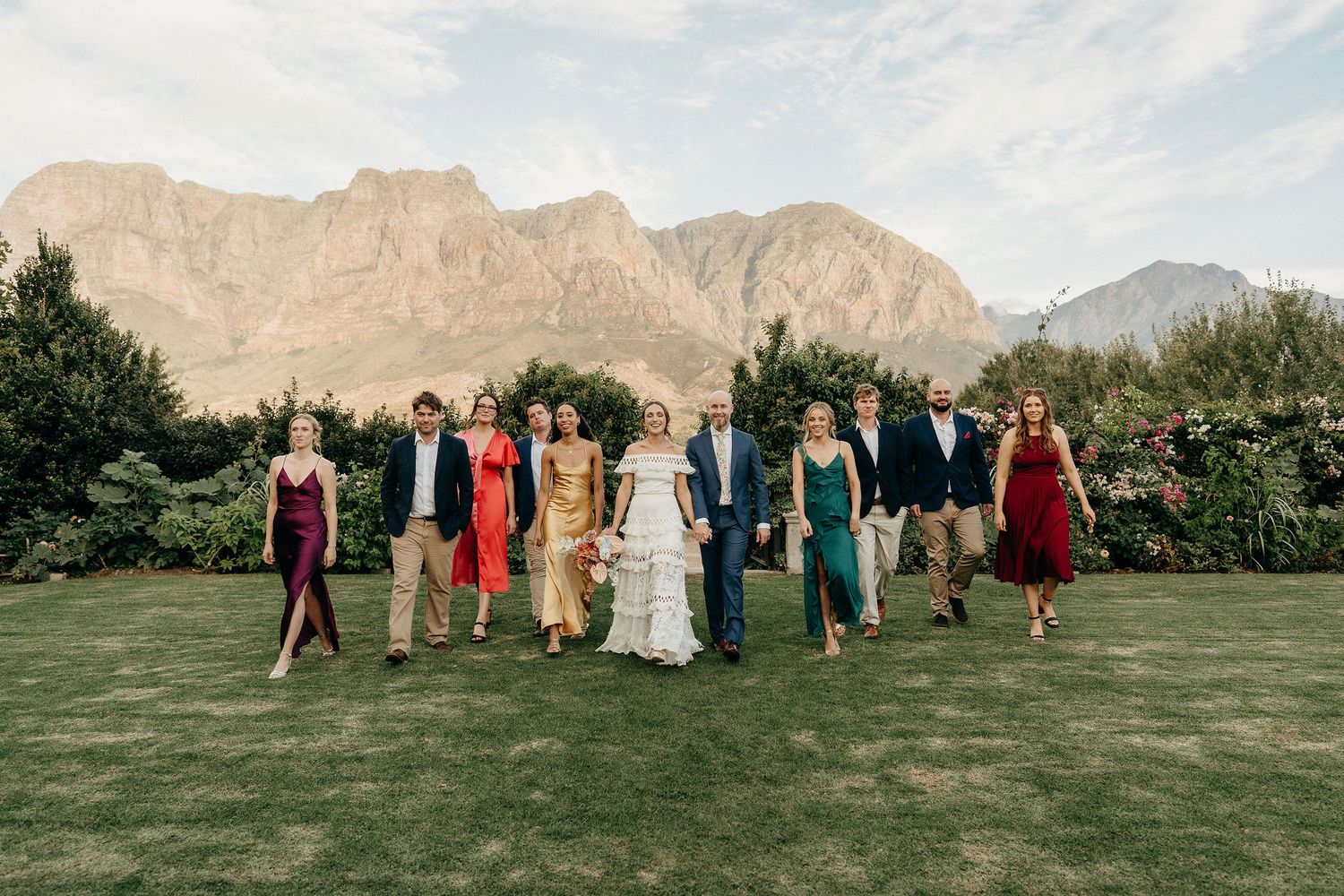 A group of wedding guests stand in formal attire on a green lawn with dramatic mountain peaks in the background.