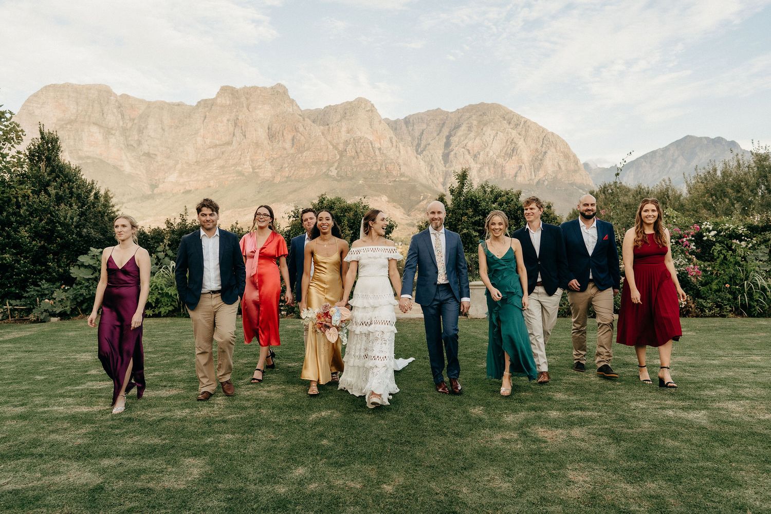 Wedding party walks together on grass with majestic desert mountains rising in the background during sunset.
