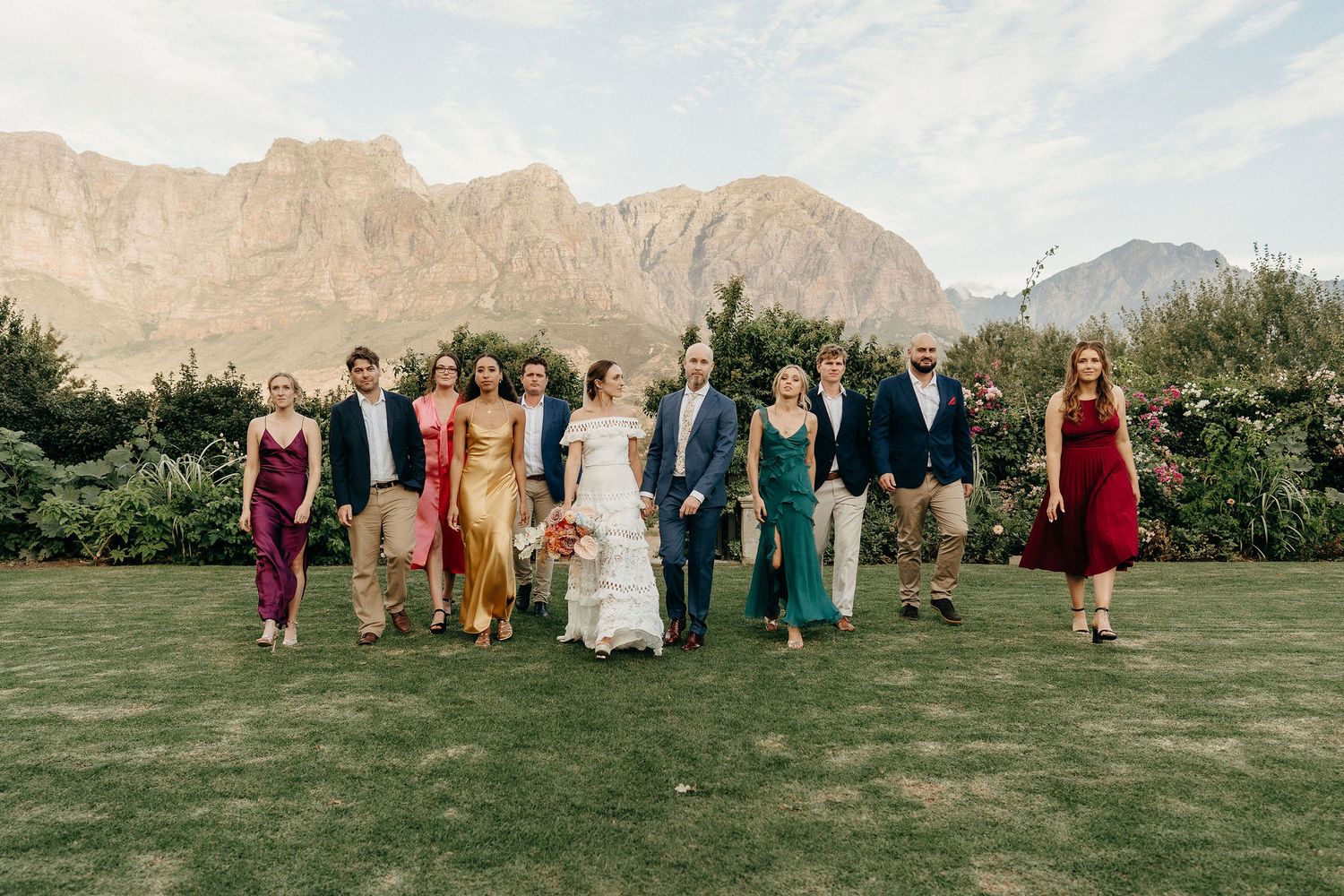 A wedding party in colorful dresses and suits poses on a lawn with dramatic desert mountains in the background.
