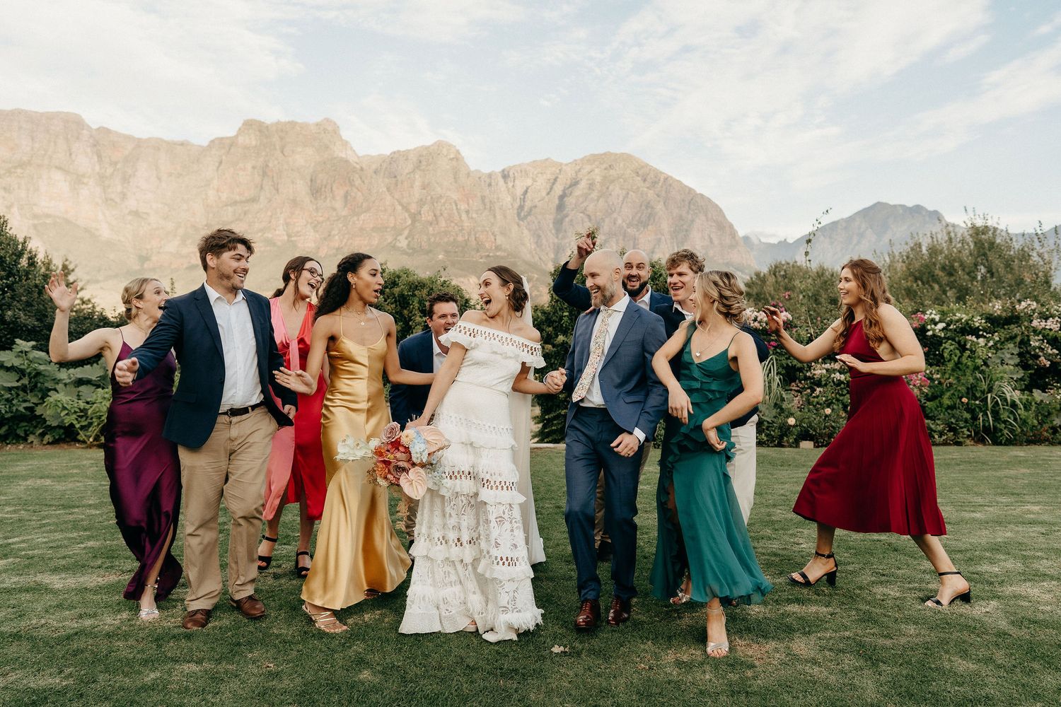 Wedding party in colorful formal attire poses outdoors against a backdrop of majestic mountains and green lawn.