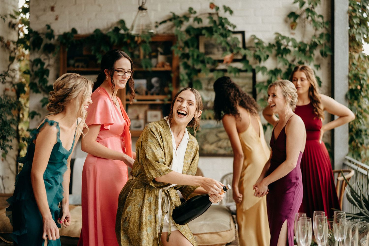 A group of friends in colorful formal dresses share a joyful moment together on a porch with ivy-covered walls.