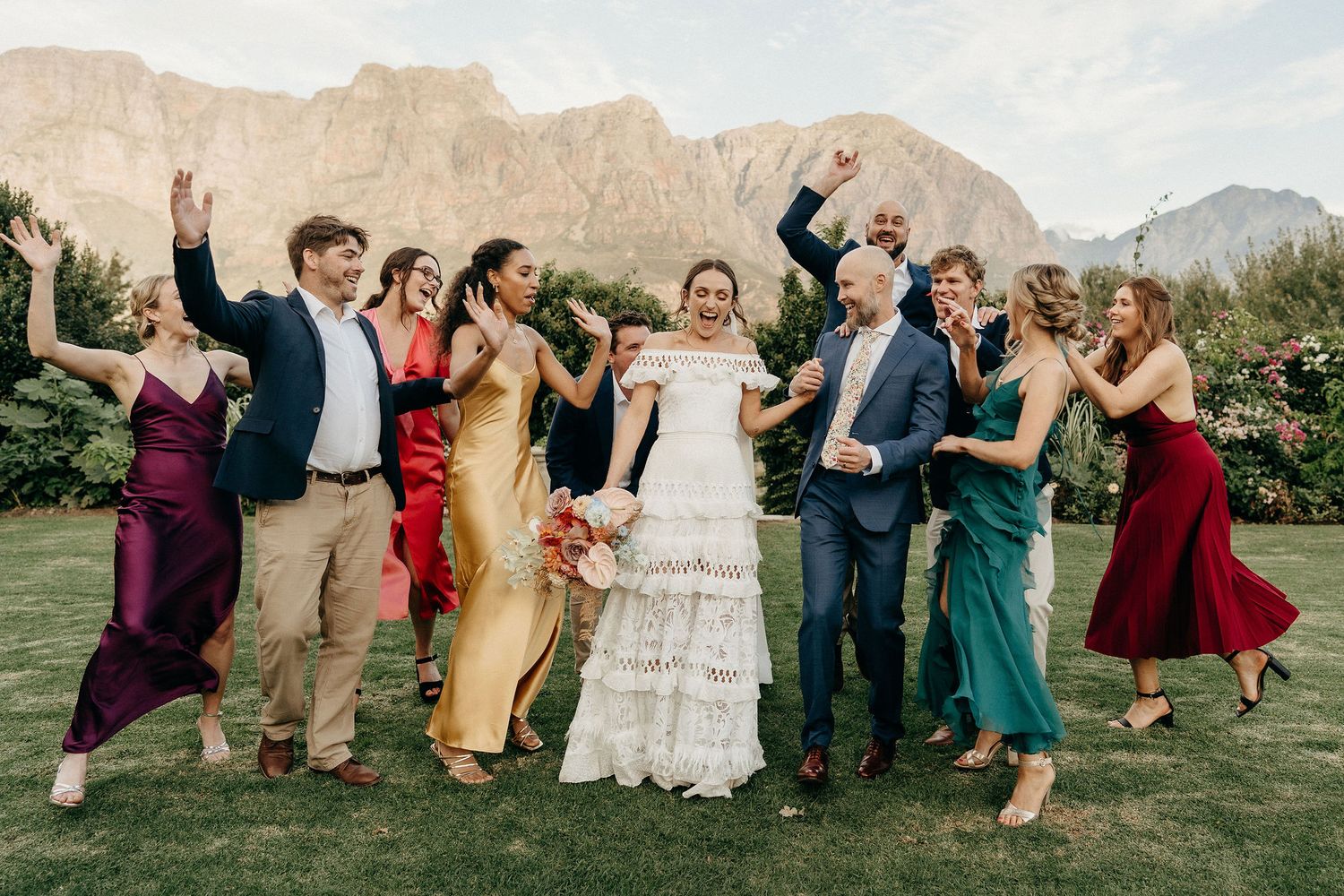 Wedding party celebrates with raised arms against mountain backdrop at outdoor venue with green lawn and desert scenery.