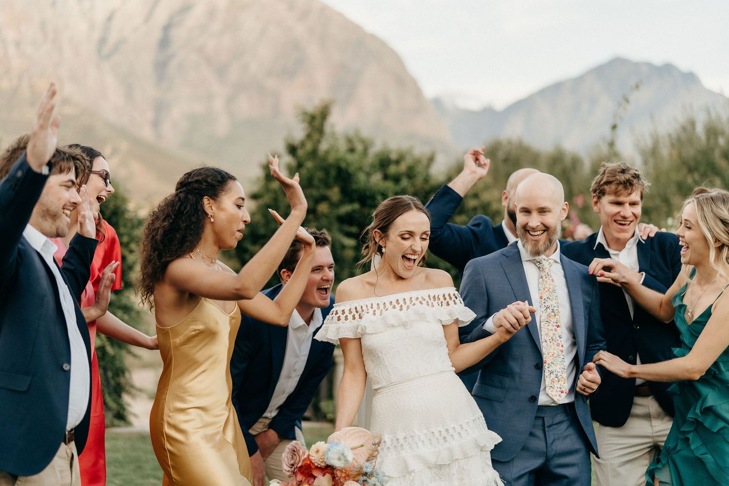 Wedding celebration with people dancing together outdoors against a scenic mountain backdrop on a sunny day.