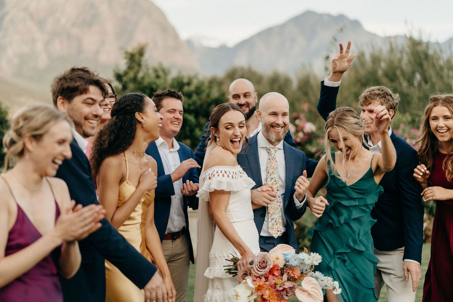 A wedding party celebrates outdoors with mountains in the background on a sunny day.