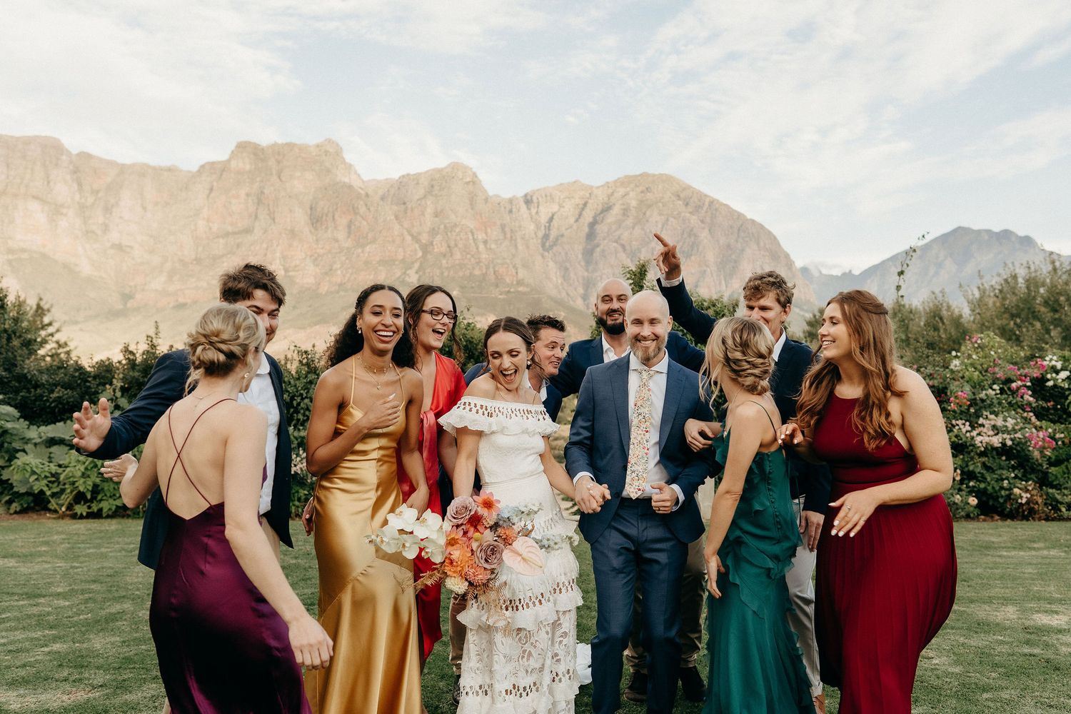 Wedding party in elegant dresses poses together on a lawn with majestic mountain peaks in the background.