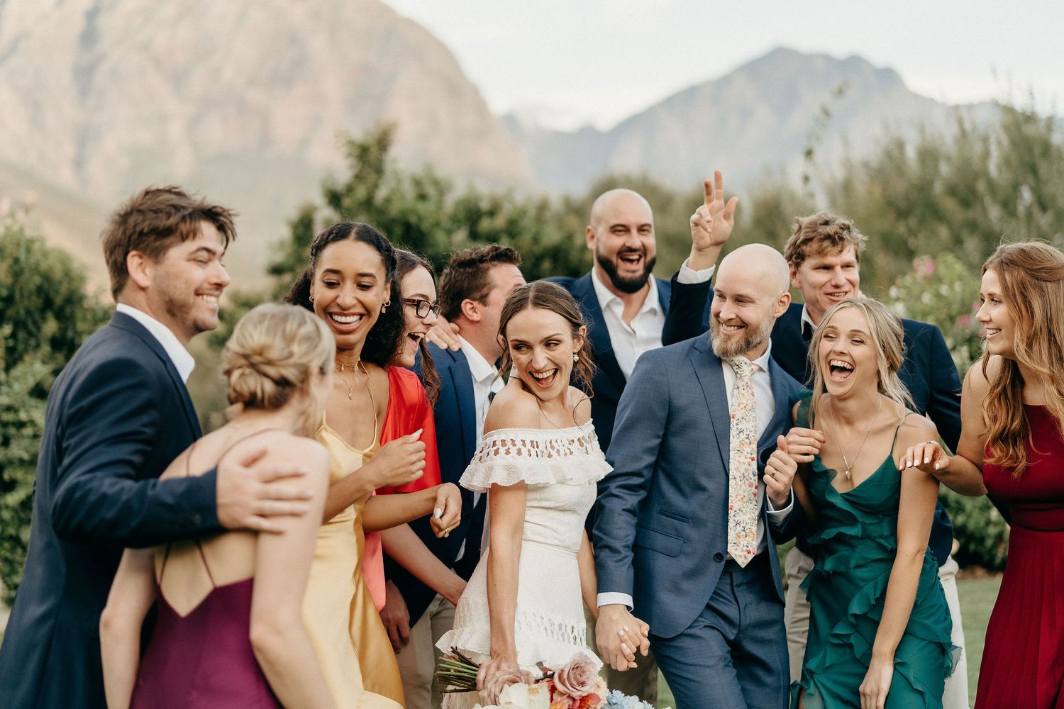 A joyful wedding party laughs together outdoors against a backdrop of mountain scenery.