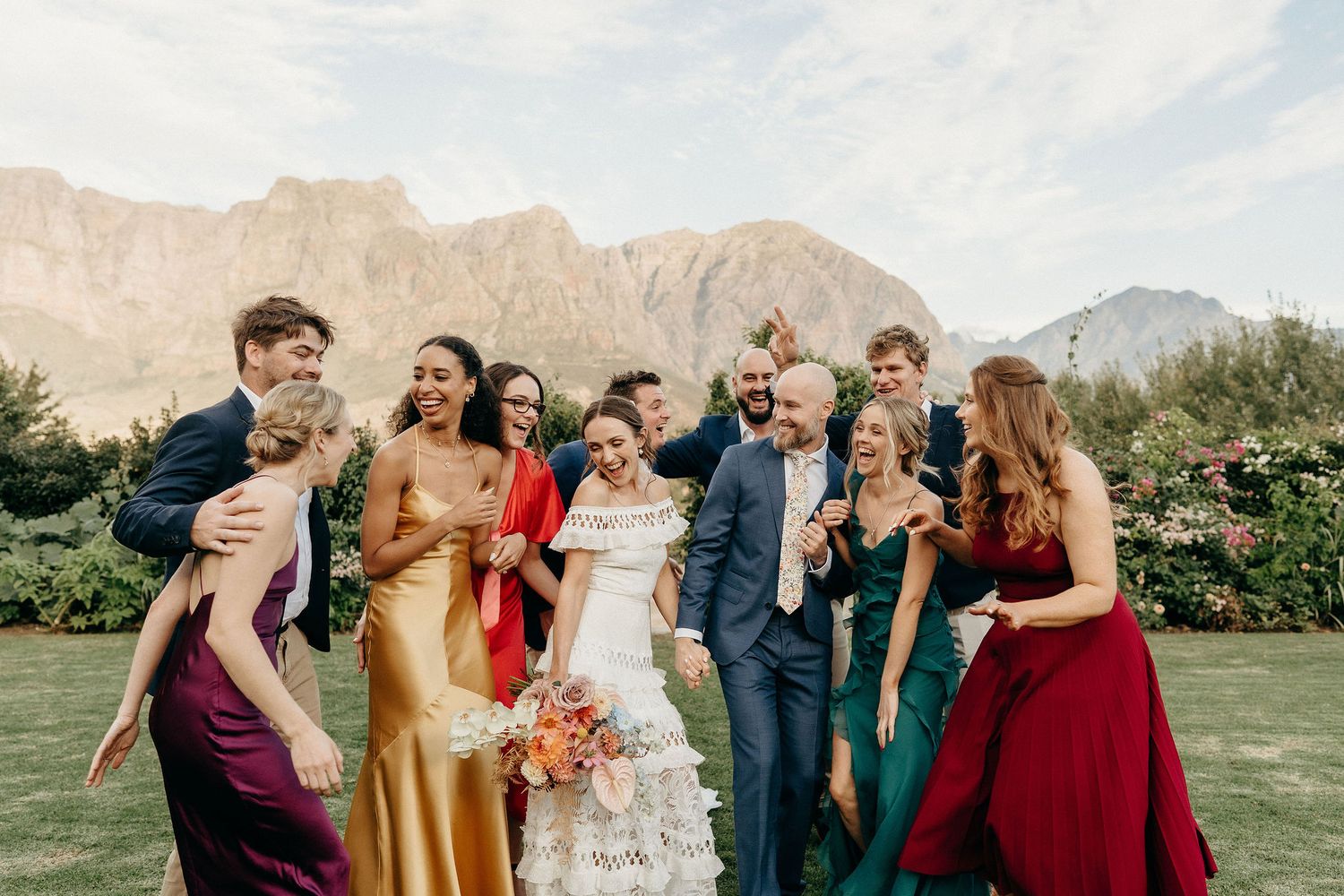 Wedding party laughs together on a grassy lawn with dramatic mountain peaks rising in the background.