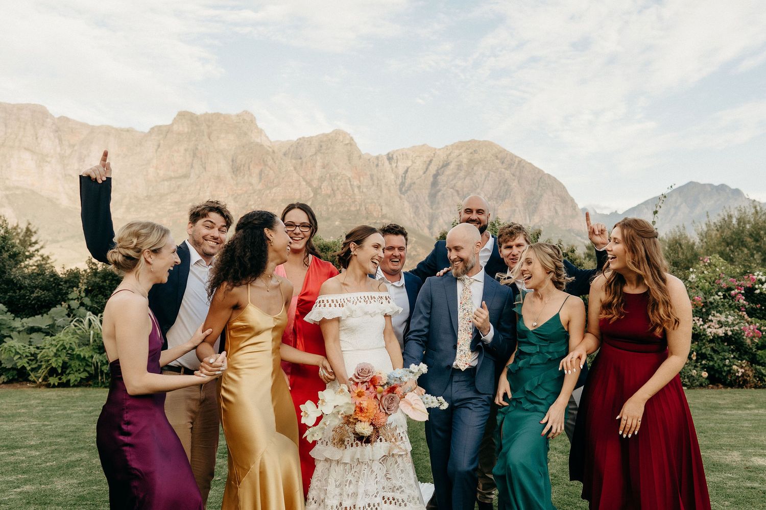Wedding party in colorful formal attire poses together on lawn with majestic mountain peaks in background.