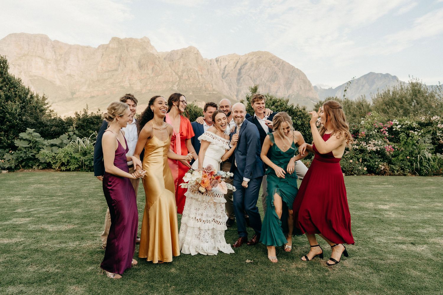 A wedding party in colorful formal attire stands on a green lawn with majestic mountains in the background.