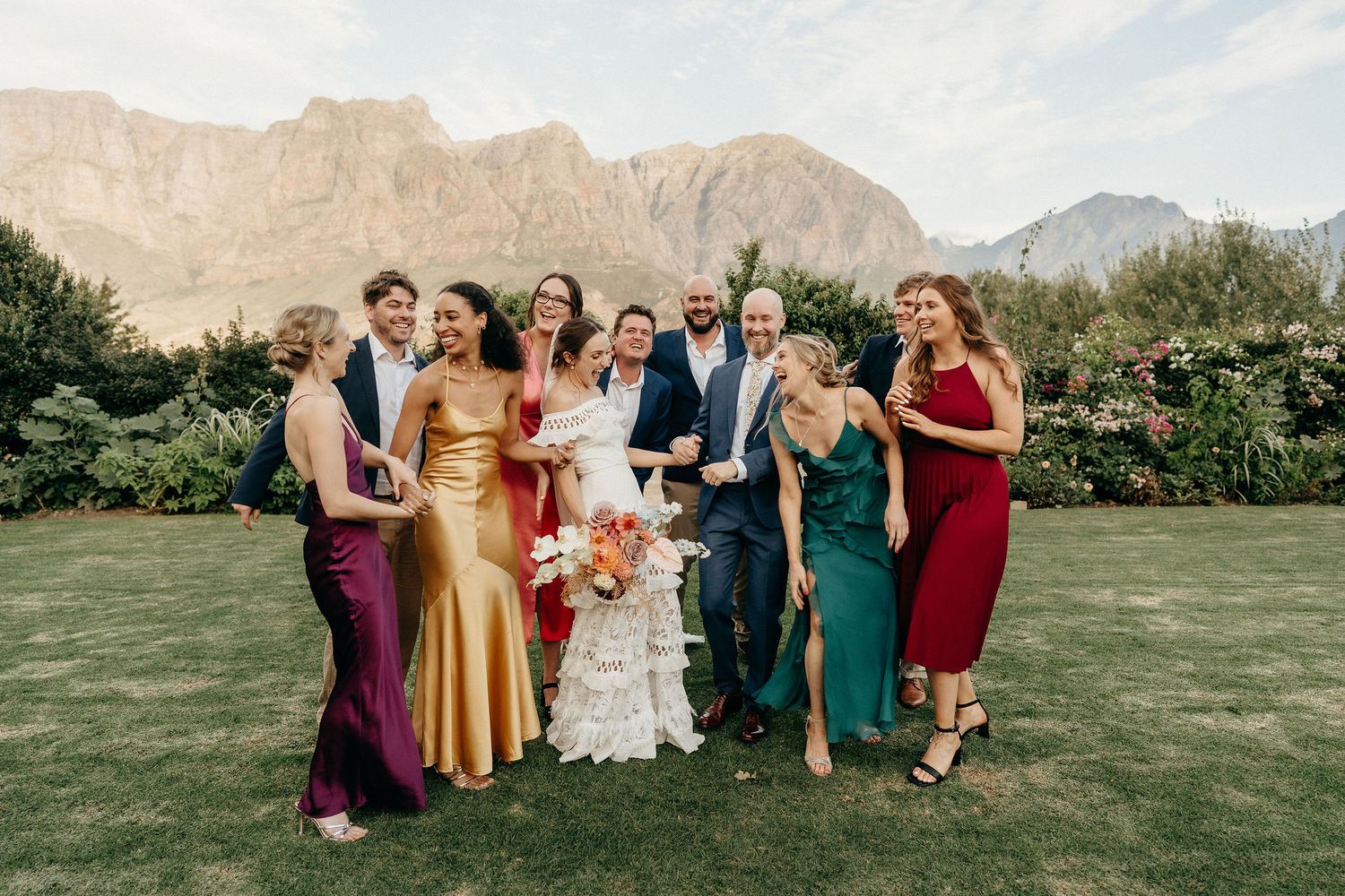 A wedding party in jewel-toned dresses poses together on a lawn with dramatic mountain peaks in the background.