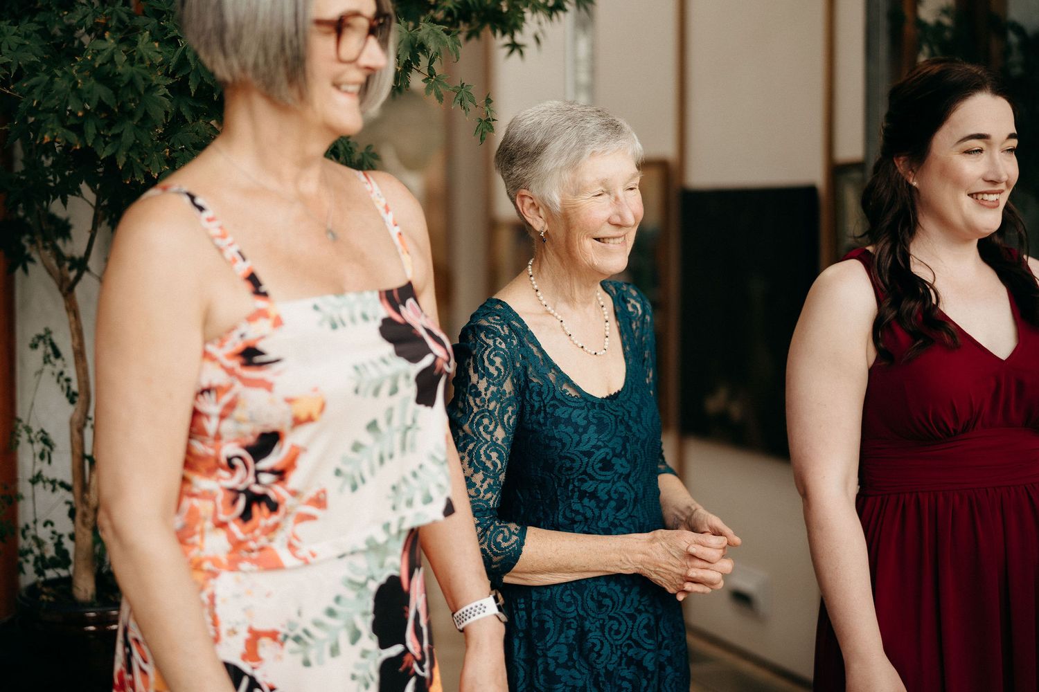 Three women share joyful laughter together while standing in semi-formal attire at an indoor gathering.