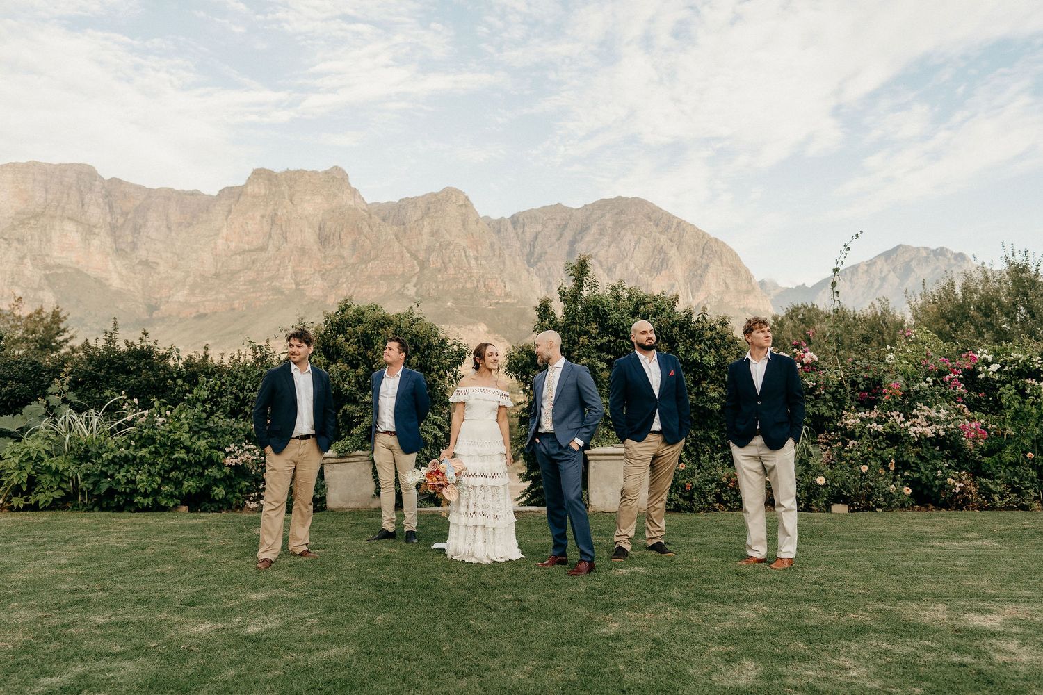 Wedding party poses on lawn with dramatic mountain backdrop in classic navy and khaki attire during outdoor celebration.