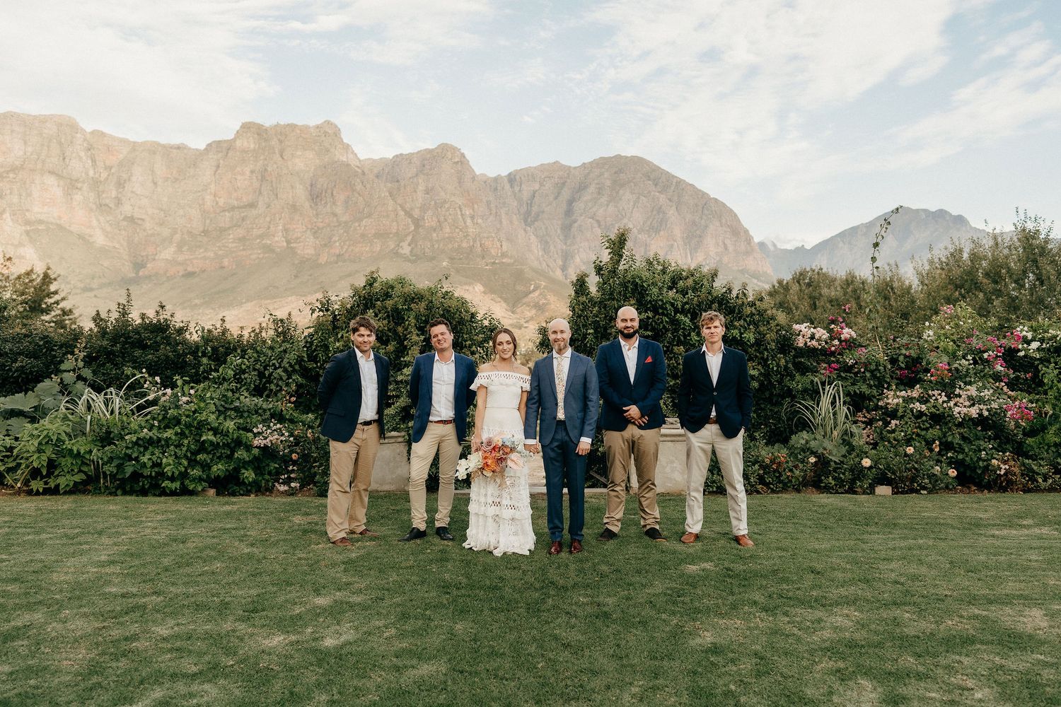 Wedding party in formal attire poses on green lawn with majestic mountain backdrop and lush garden landscape.