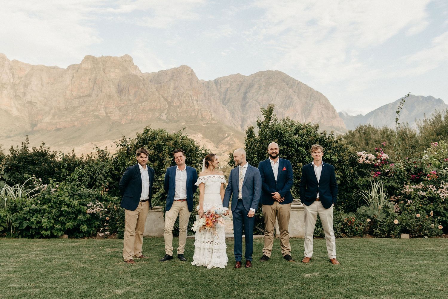 Wedding party poses on a green lawn with dramatic mountain peaks and lush foliage as their backdrop.