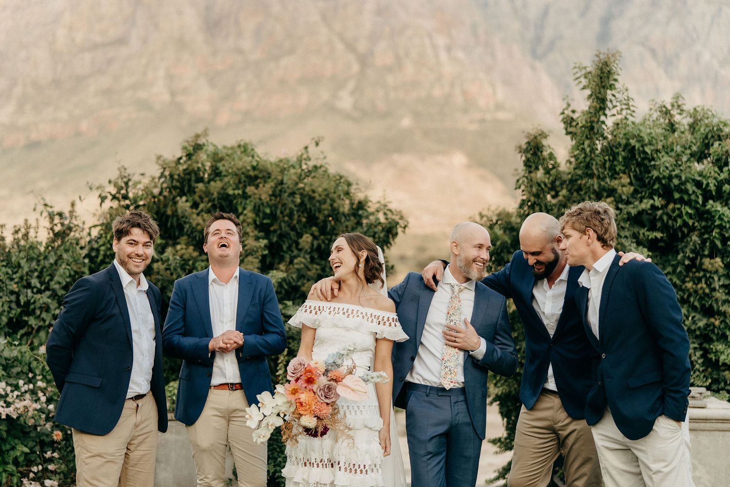 Candid wedding party laughs together on scenic lawn with desert mountains and greenery in background.