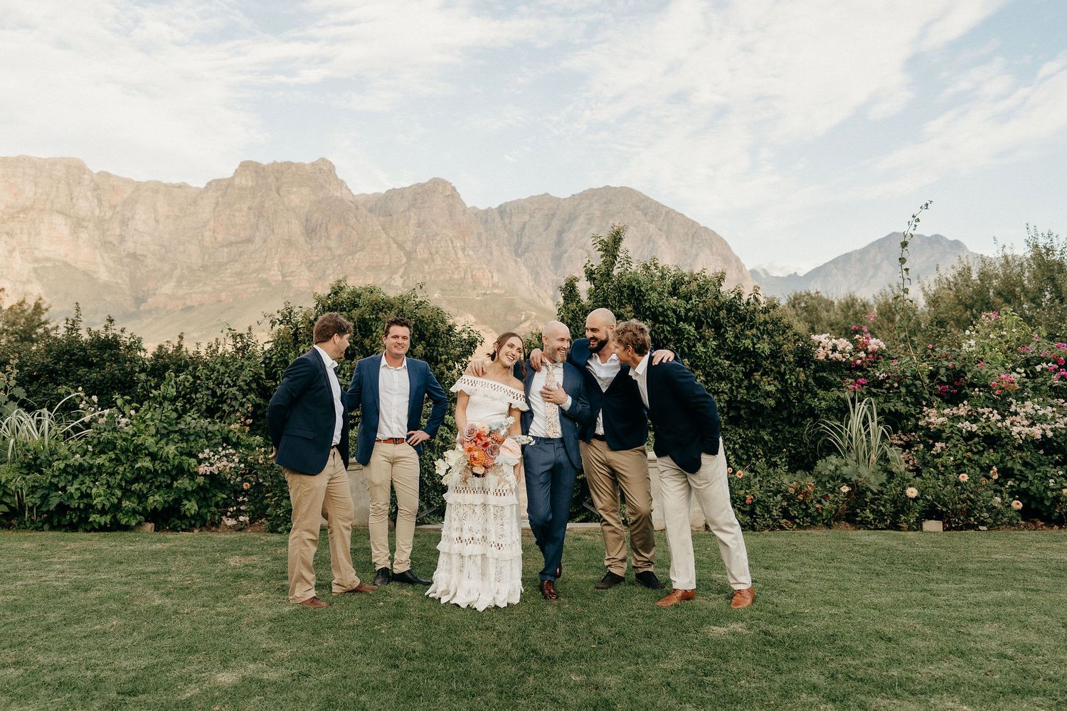 Wedding group poses together on a manicured lawn with dramatic mountain landscape and greenery in the background.