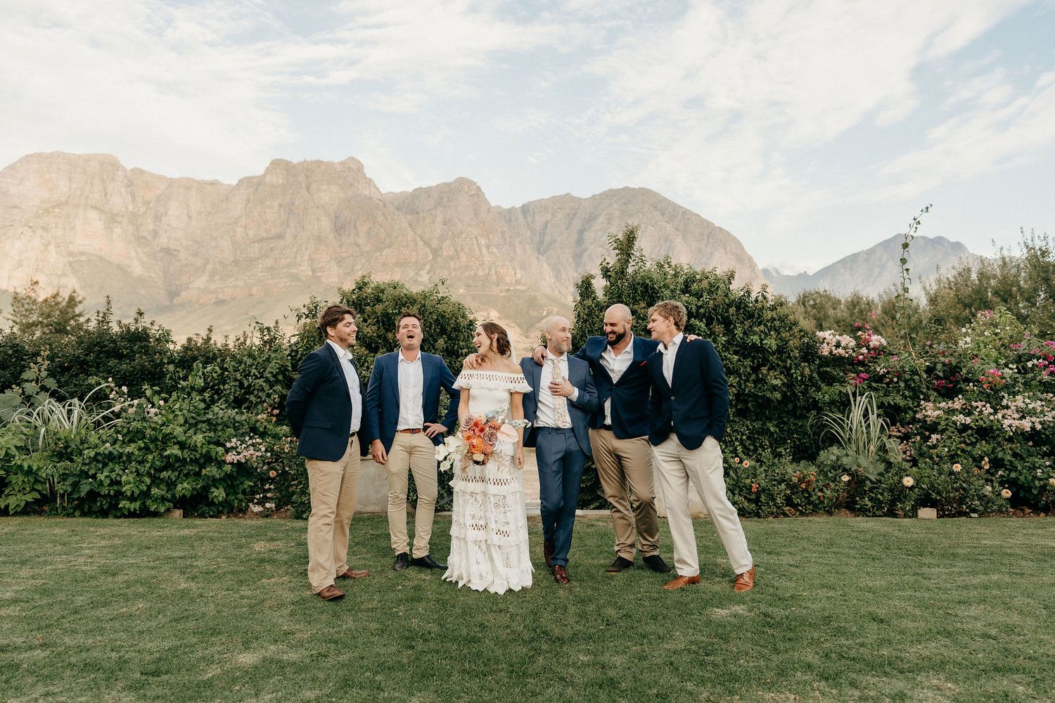 Group poses on green lawn with lush garden and scenic mountain backdrop in casual formal attire.