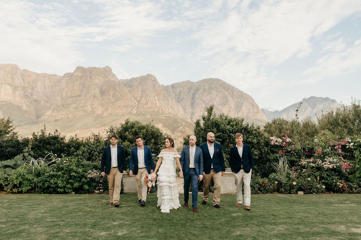 Wedding party poses on green lawn with dramatic mountain peaks rising in background at winery venue.