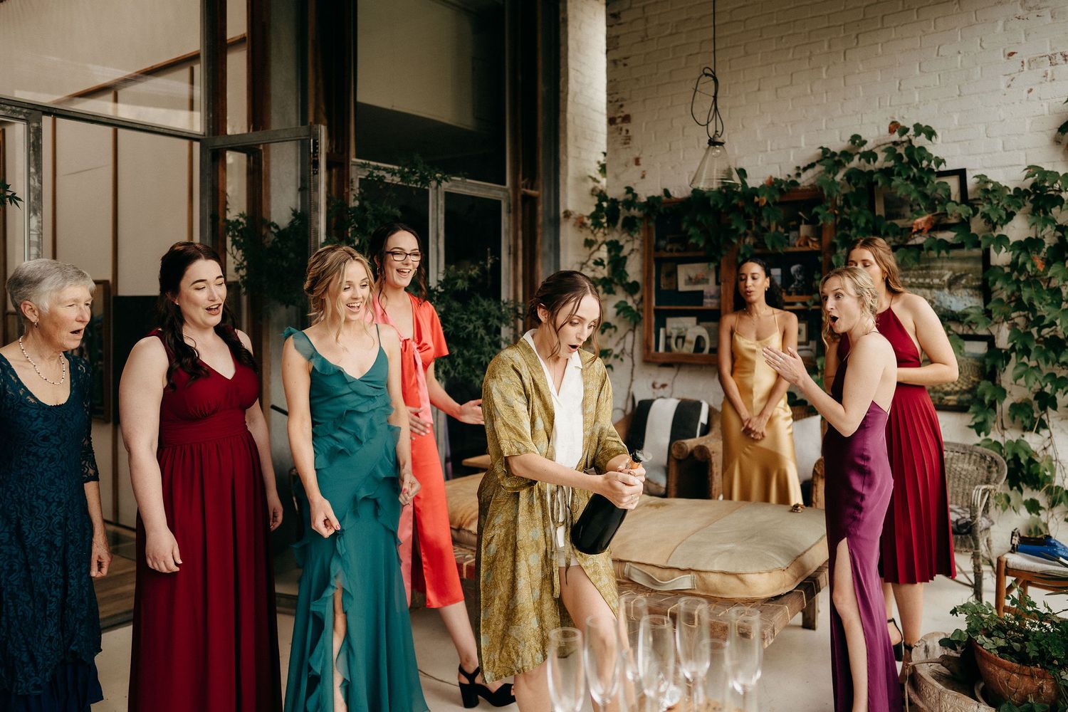 A group in elegant dresses and robes gather around a table during a celebratory event in a bright indoor space with plants.