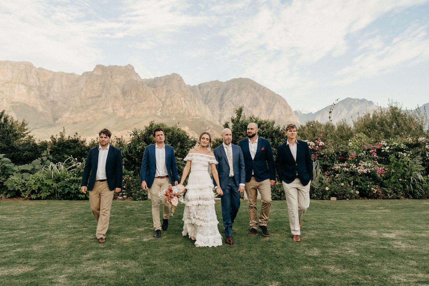 Wedding group walks across green lawn with stunning mountain scenery and garden landscape behind them.