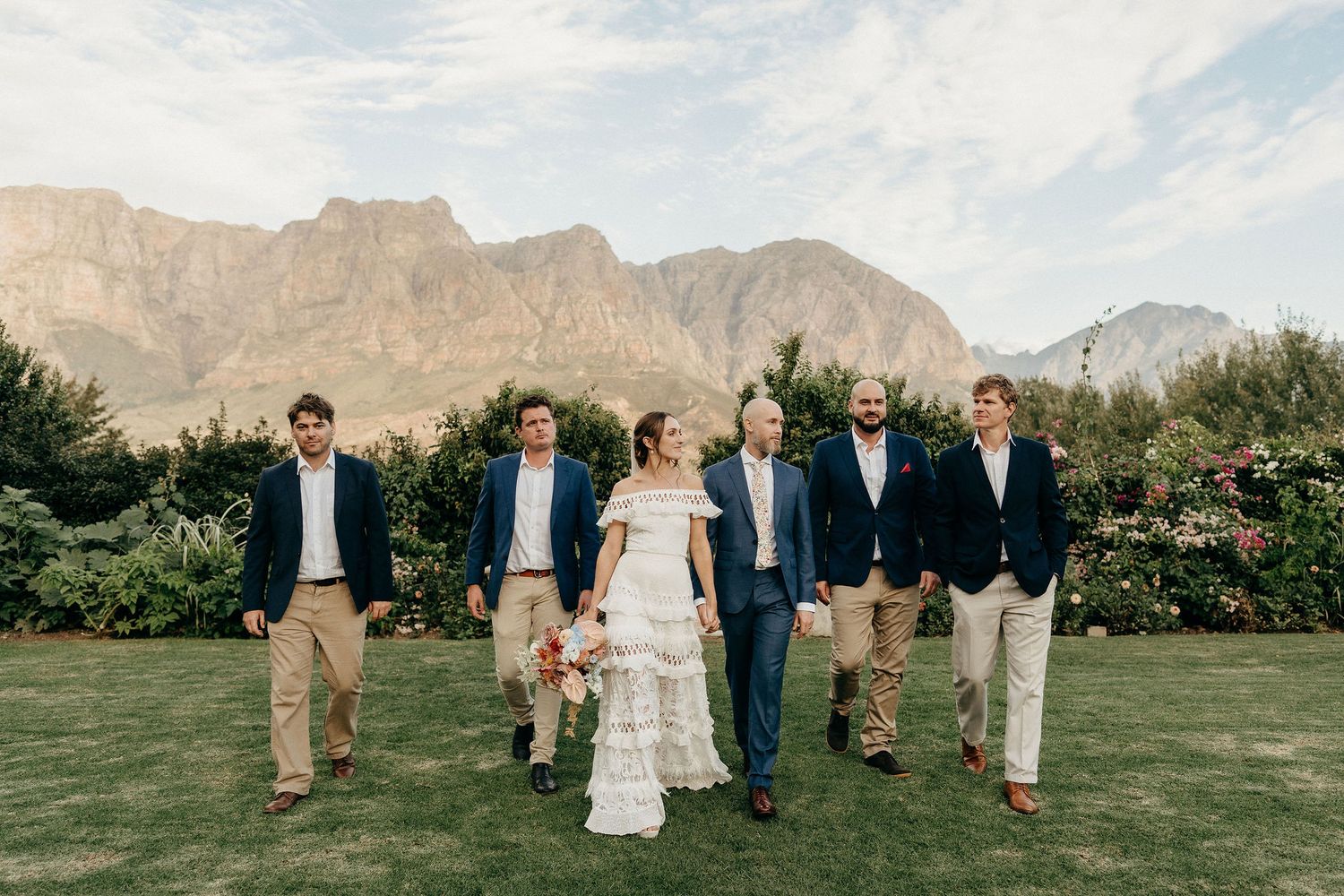Wedding party in Navy and khaki walking across manicured lawn with dramatic mountain backdrop in South Africa.
