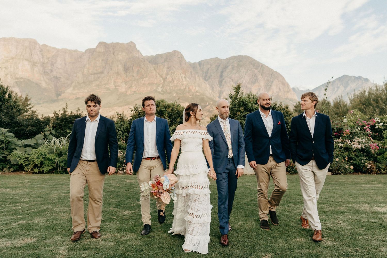 Wedding party walks together across lawn with majestic mountain peaks rising in background on sunny day.