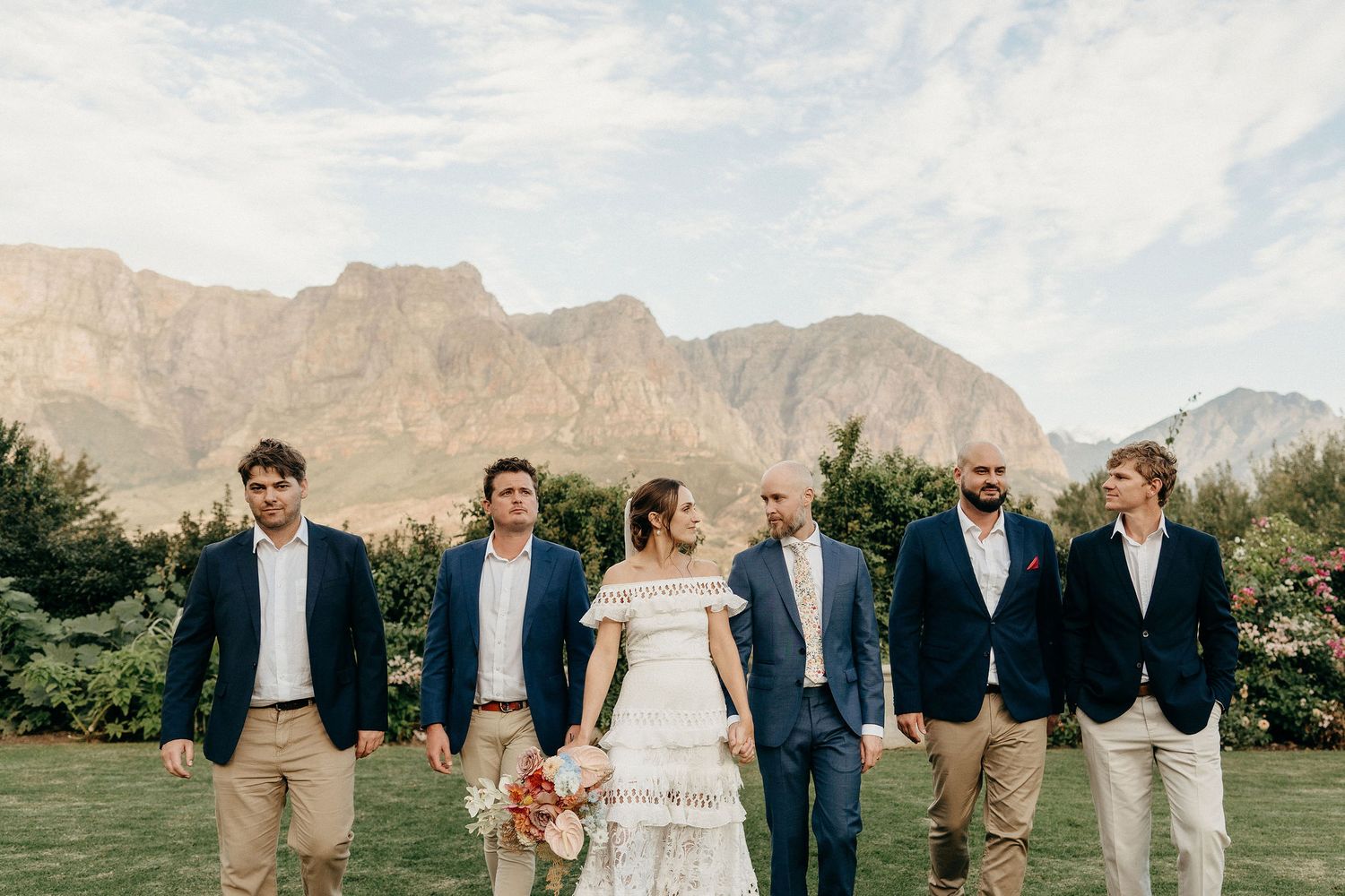 Wedding party stands together against backdrop of dramatic mountains at a winery in South Africa.