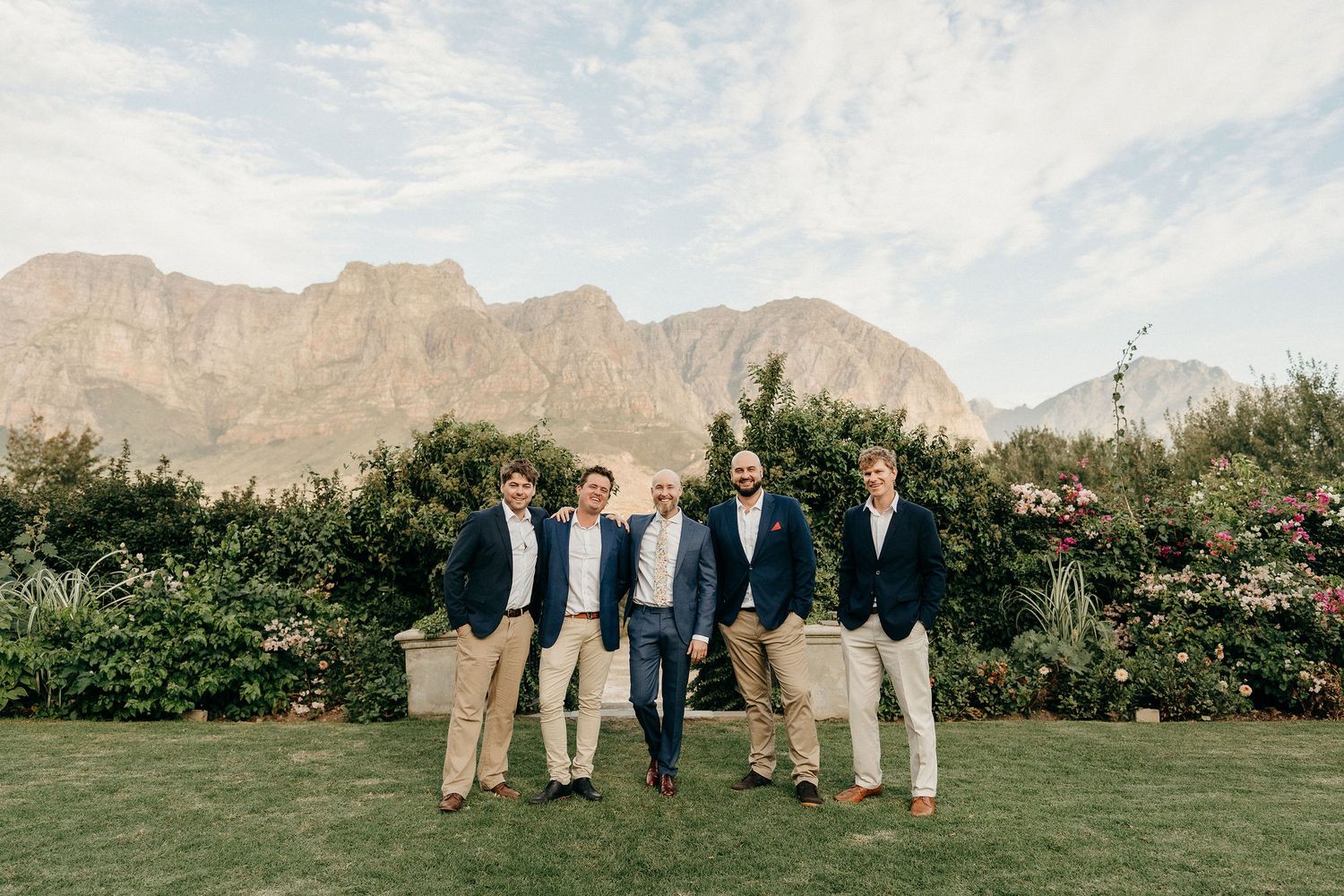 Group of people in formal attire standing on a lawn with majestic mountain peaks and greenery in the background.