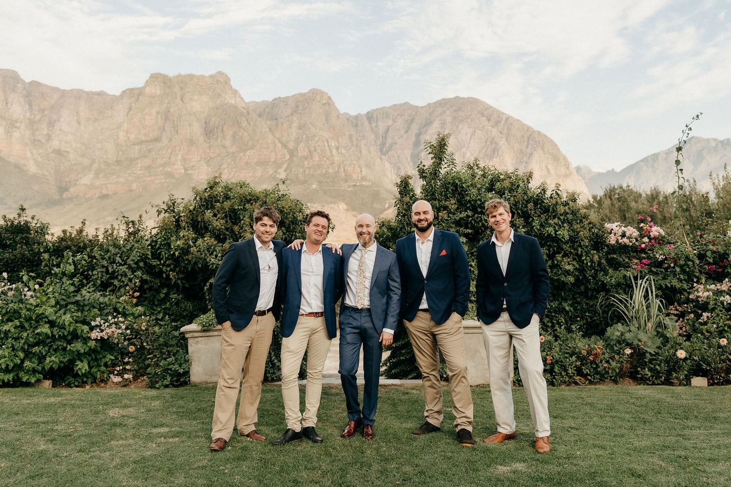 A group of five people in navy blazers and khaki pants pose together on a lawn with mountains in the background.