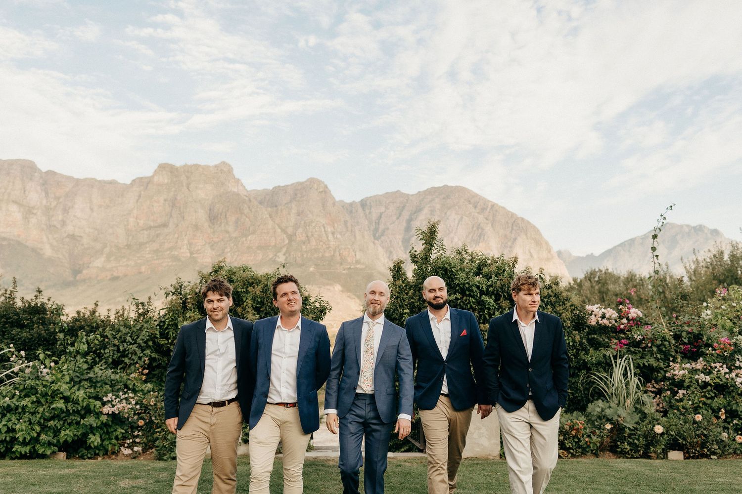 A group of groomsmen in khaki pants and navy jackets stand in front of a mountain landscape and garden.