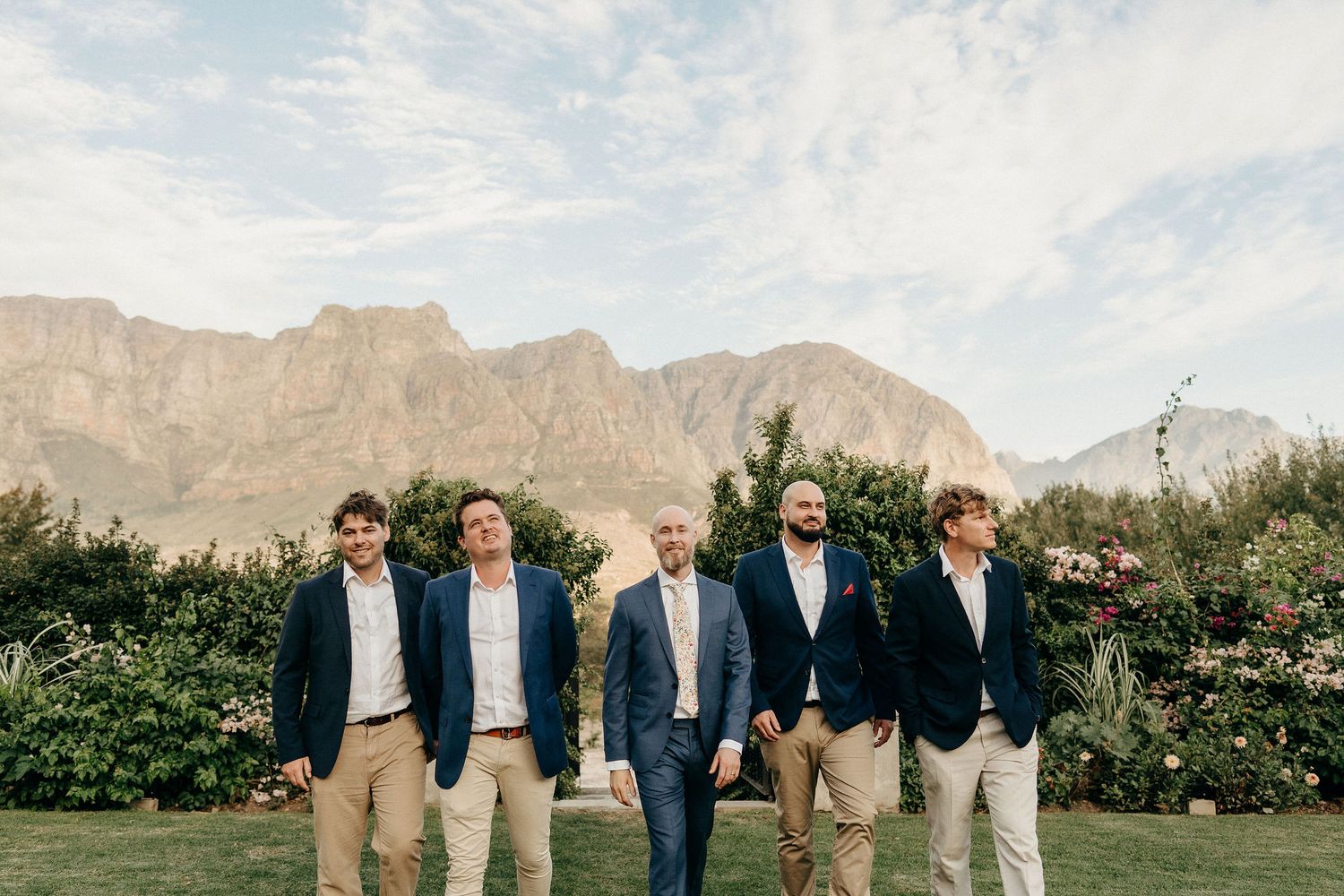 Five groomsmen in matching navy blazers and khaki pants pose outdoors with dramatic mountain peaks in the background.