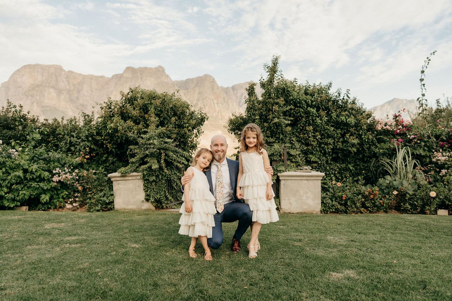 Two children in light-colored dresses stand together on a garden lawn with mountains visible in the background.