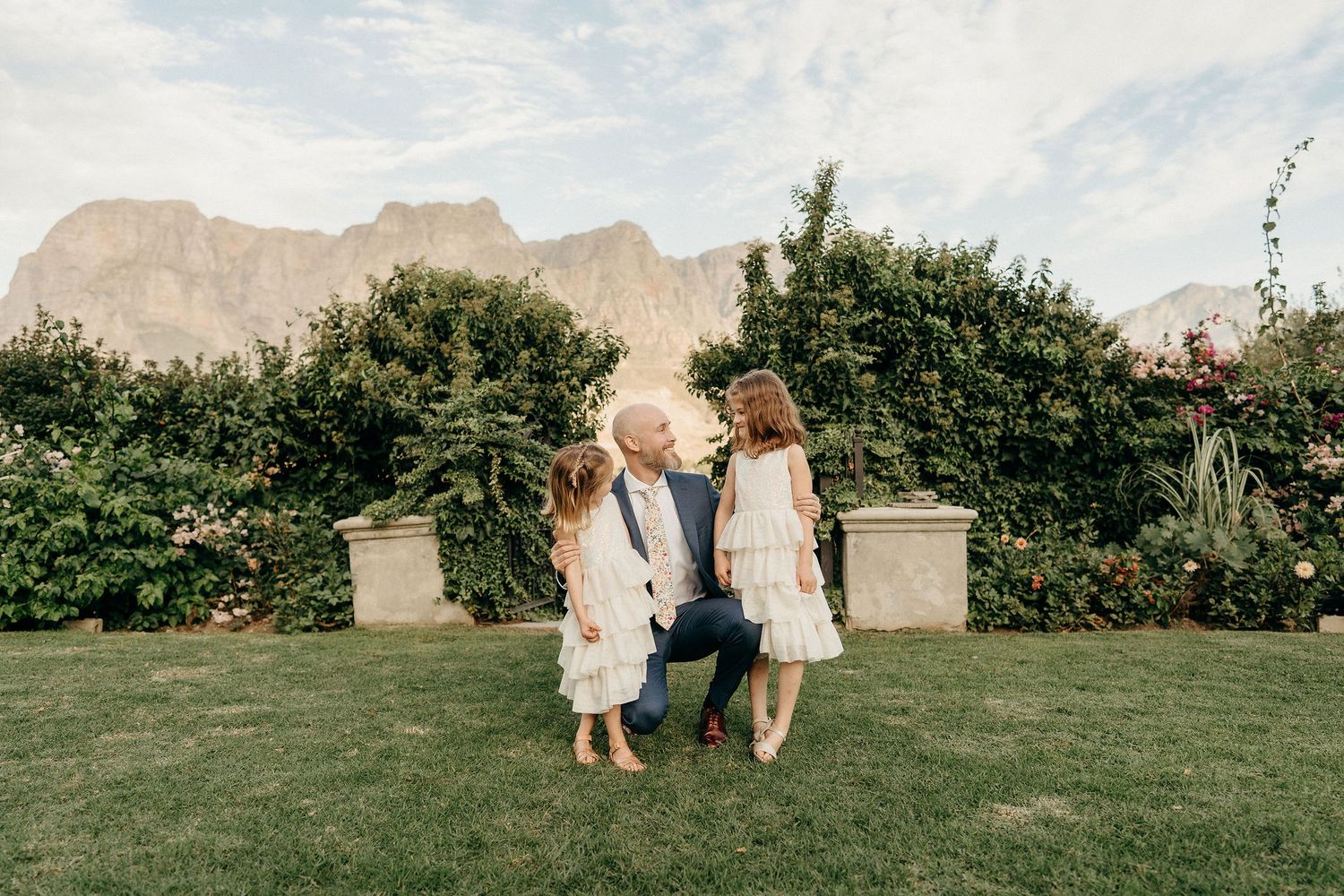 Two children in white outfits hold hands in a garden with mountain peaks visible in the background.
