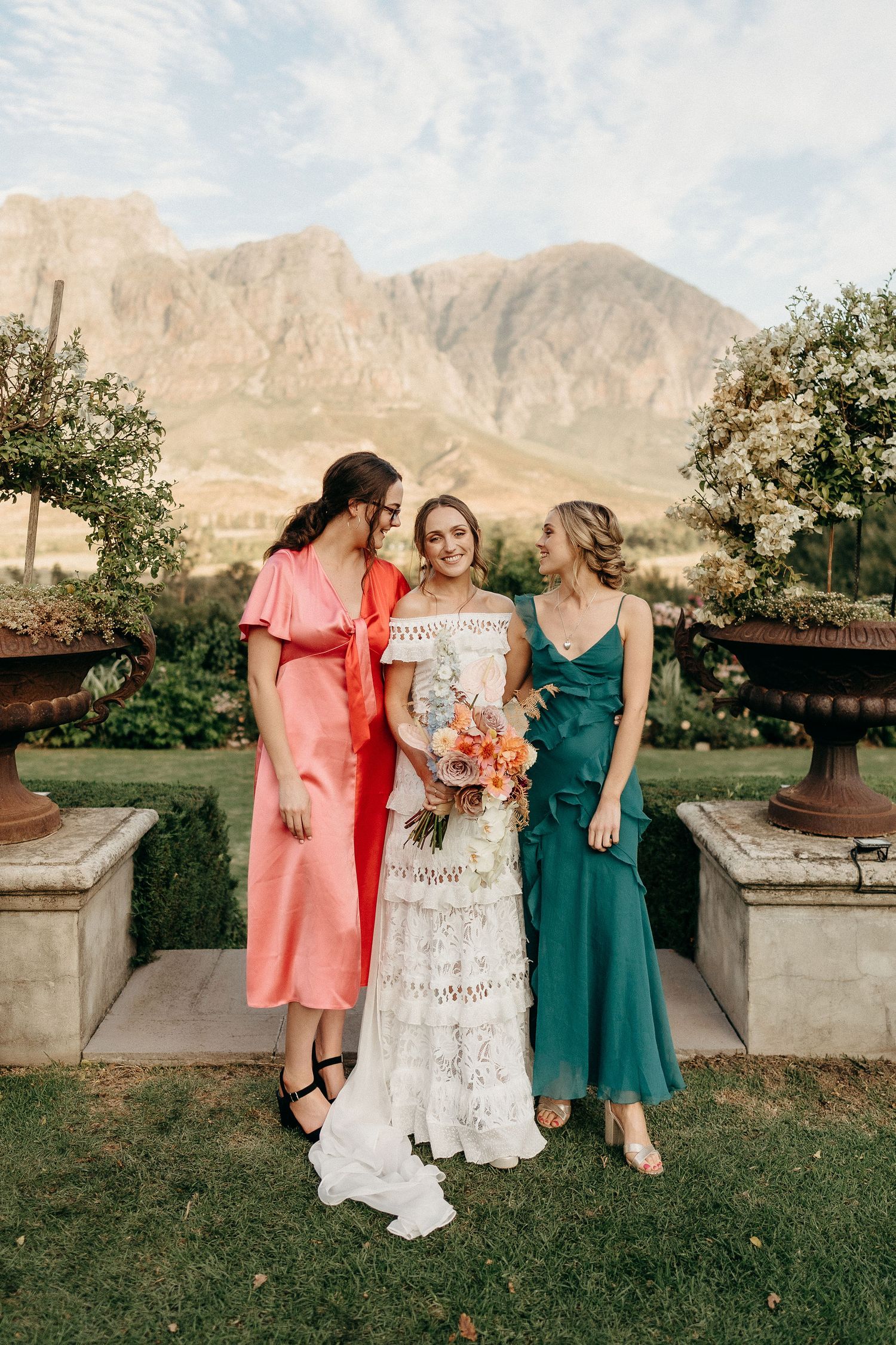 Three women in formal dresses pose together at an outdoor venue with dramatic mountain peaks in the background.