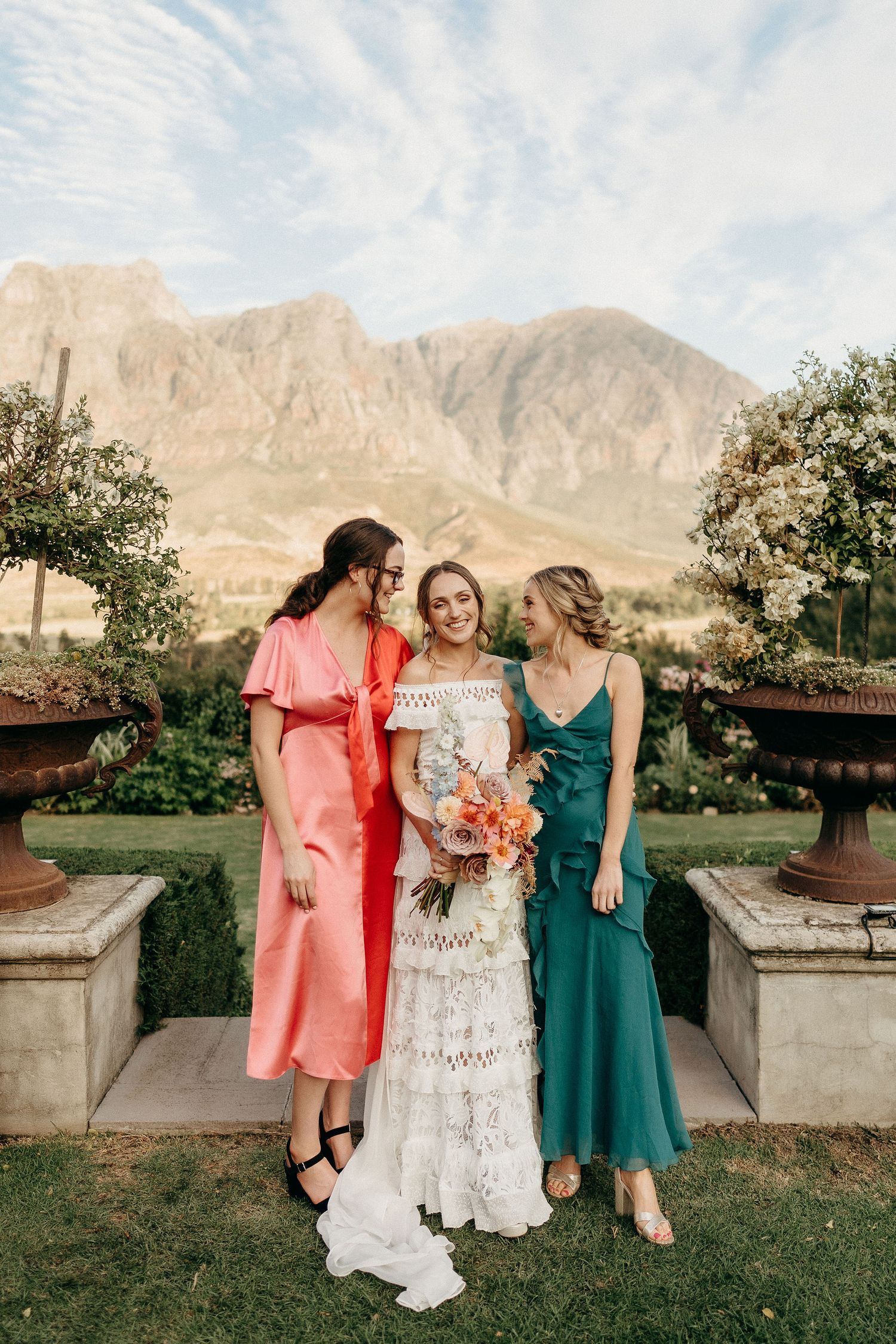 Three women in elegant dresses pose together at an outdoor venue with dramatic mountain backdrop and ornate stone planters.
