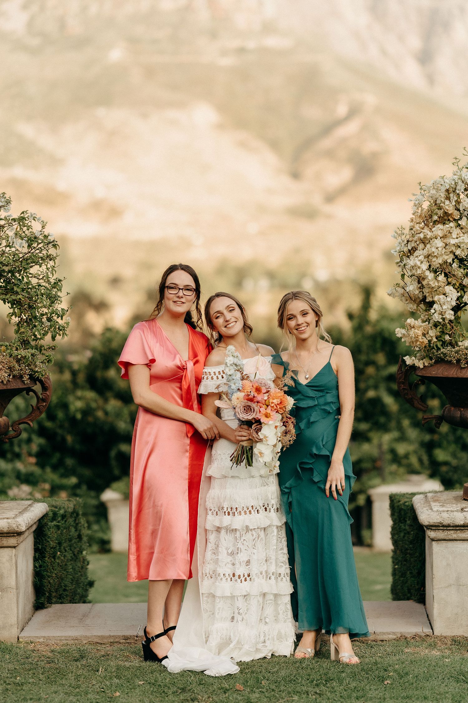 Three people in colorful dresses pose together at an outdoor garden wedding with mountains in the background.