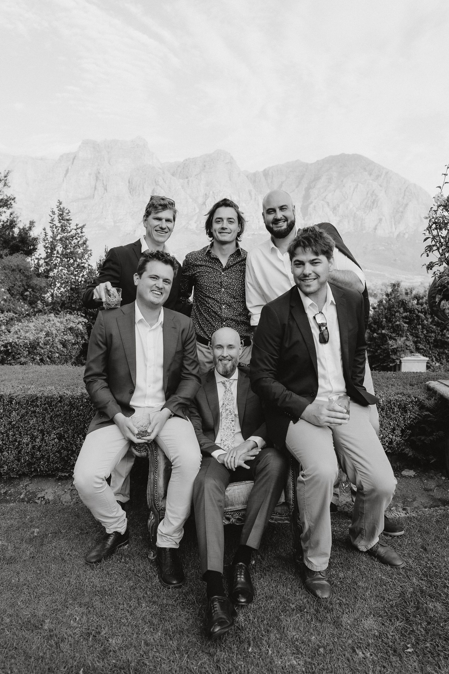 Black and white photo of six men dressed in formal attire posing together outdoors with mountain landscape backdrop.
