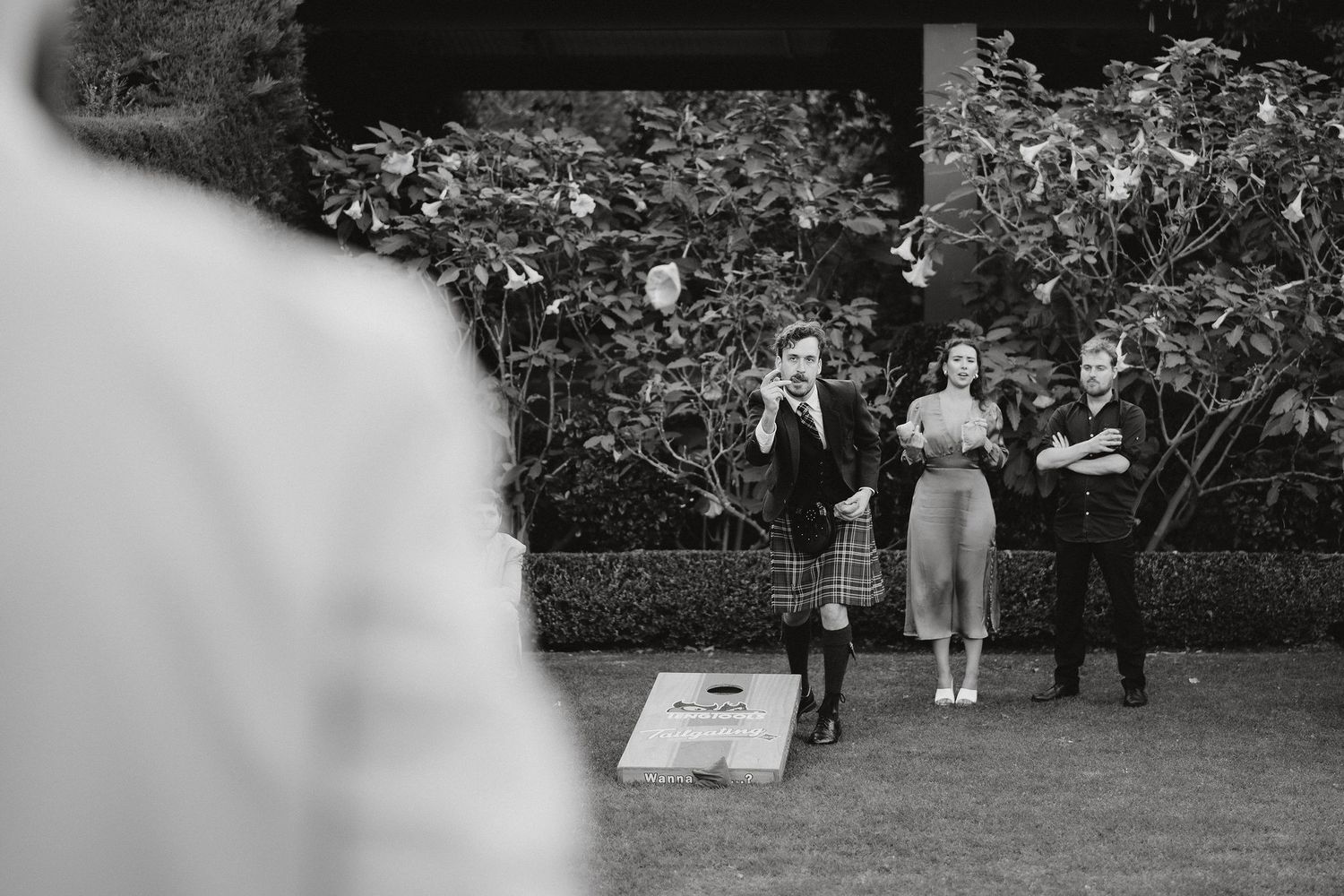 A group of people playing a game of cornhole in a backyard garden setting captured in black and white.
