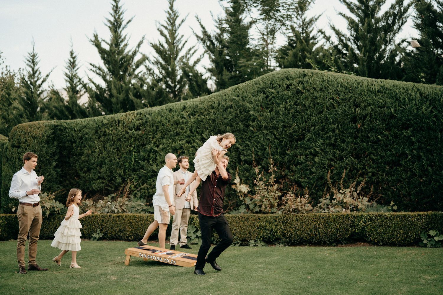 People playing and dancing on a green lawn with evergreen trees and manicured hedges in the background.
