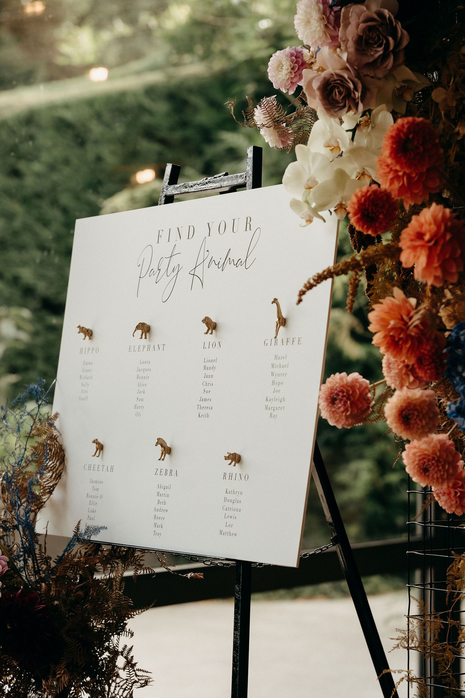 A wedding seating chart decorated with whimsical animal illustrations and floral arrangements in peach and rose tones.