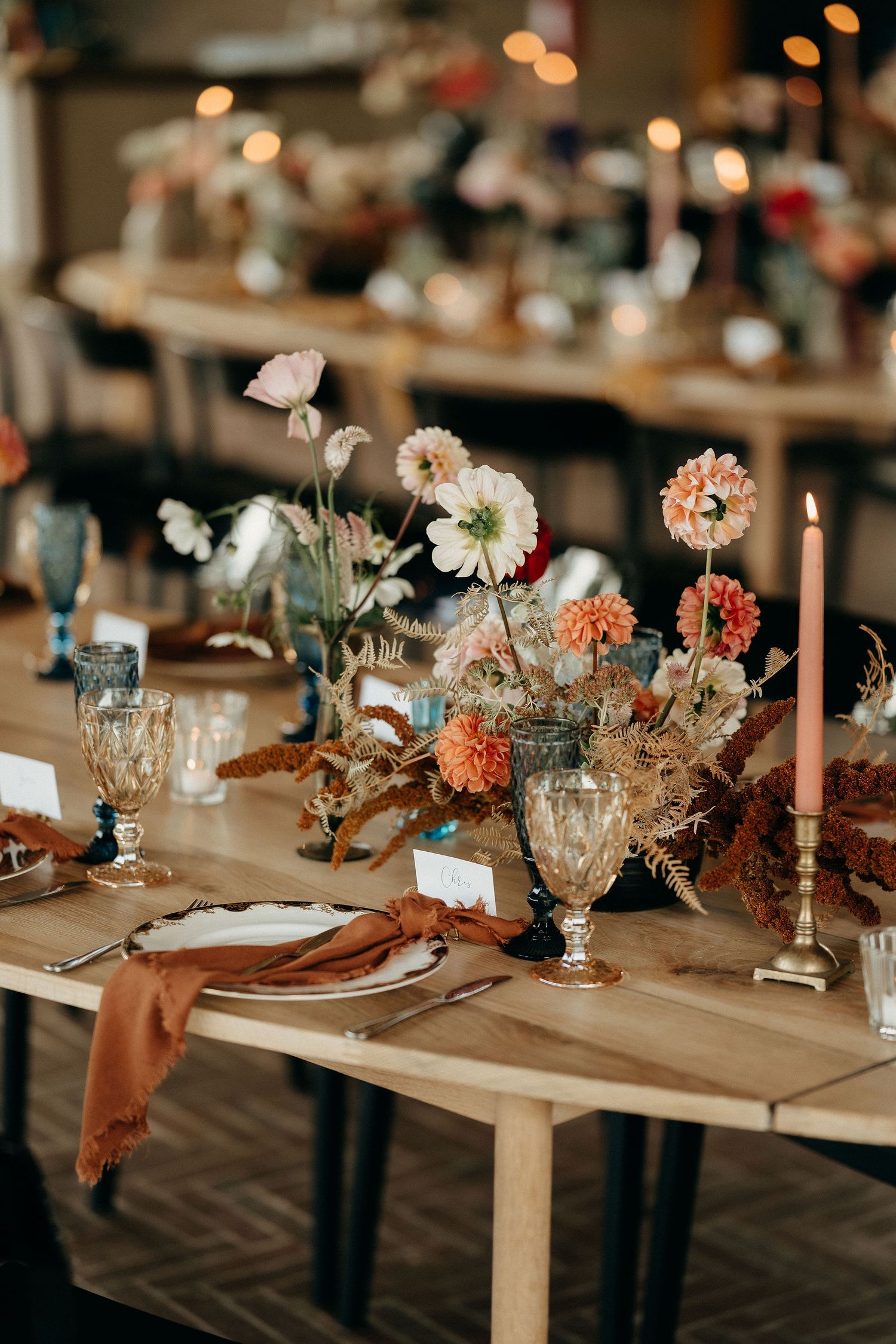 Rustic wedding table setting with peach and white flowers, vintage glassware, and terracotta napkins on wooden table.