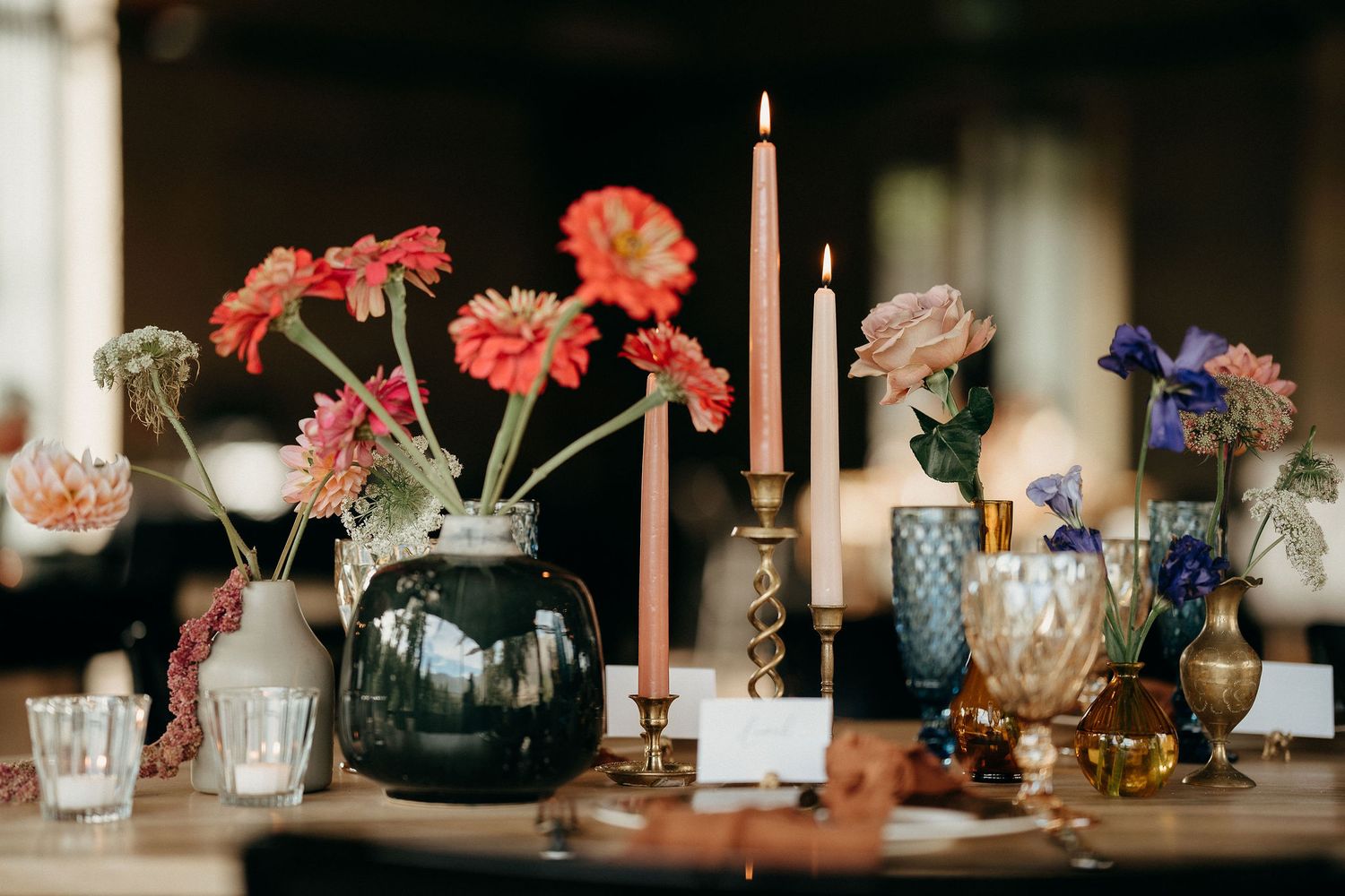 Wedding table decor with pink gerbera daisies in vases, tall pink candles, and vintage glassware on wooden surface.