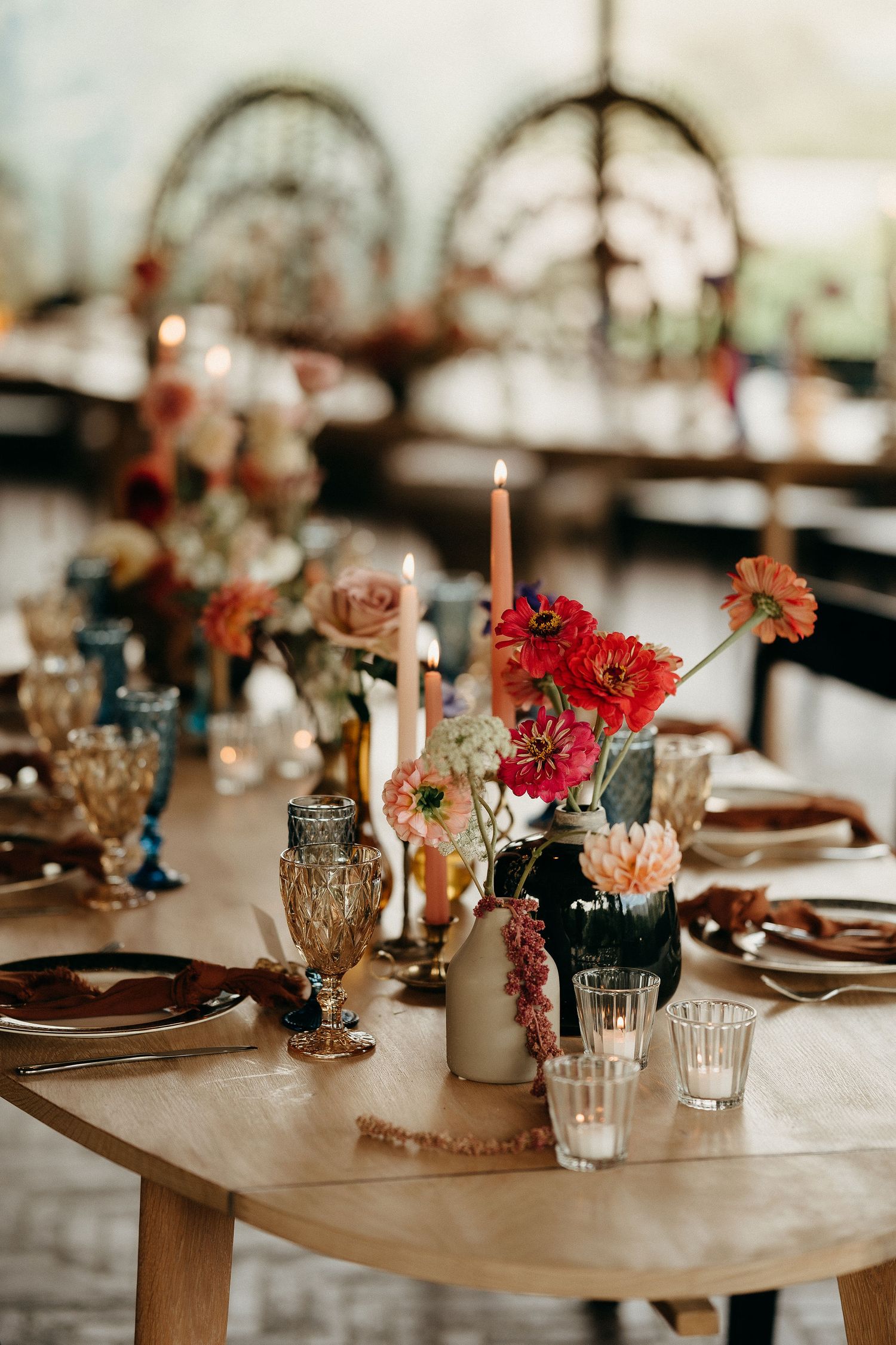 Elegant wedding table setting with red flowers, taper candles and vintage glassware on a wooden table in a rustic venue.