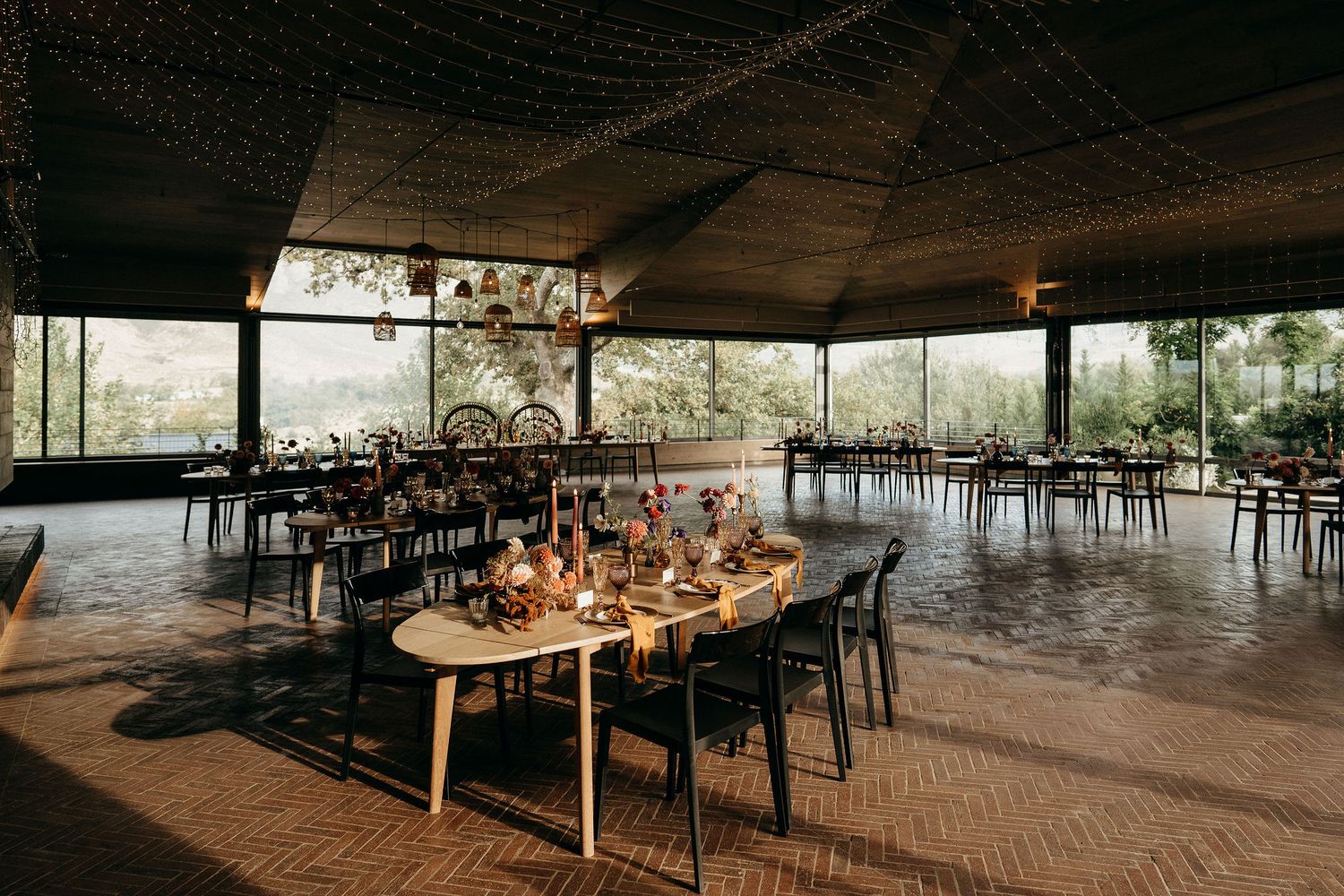 Rustic outdoor restaurant patio with wooden tables, black chairs and warm lighting under a covered pavilion structure.