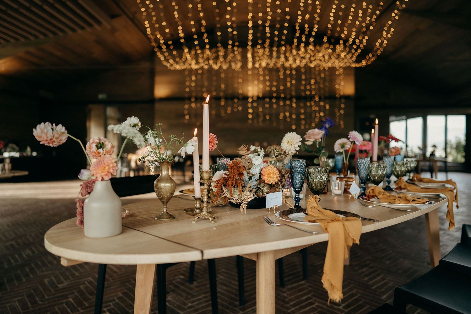 A rustic wedding reception table with delicate string lights overhead and floral centerpieces in white and coral tones.