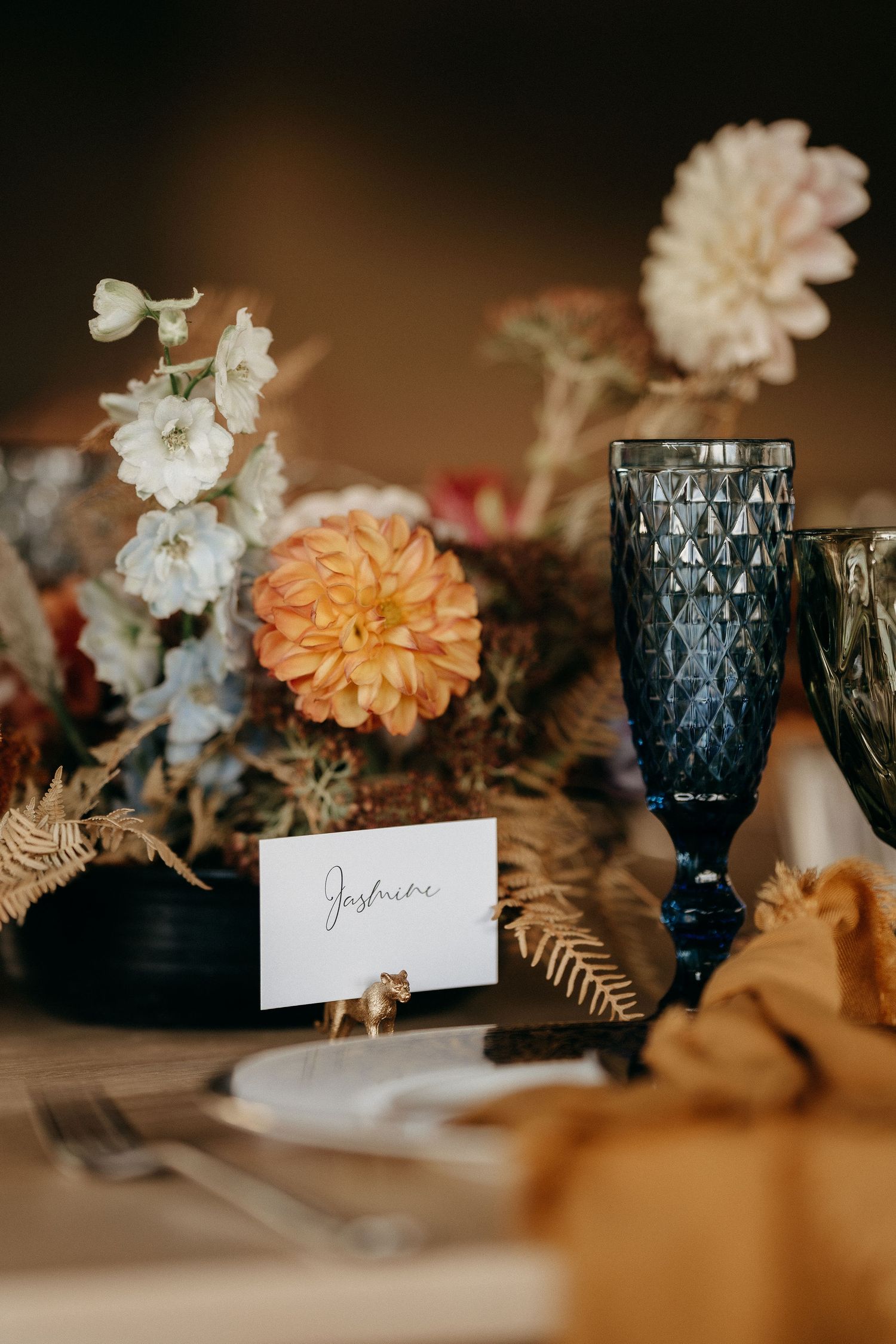 Elegant table setting with coral dahlia flowers, white blooms, navy blue glassware and a place card on a rustic table.