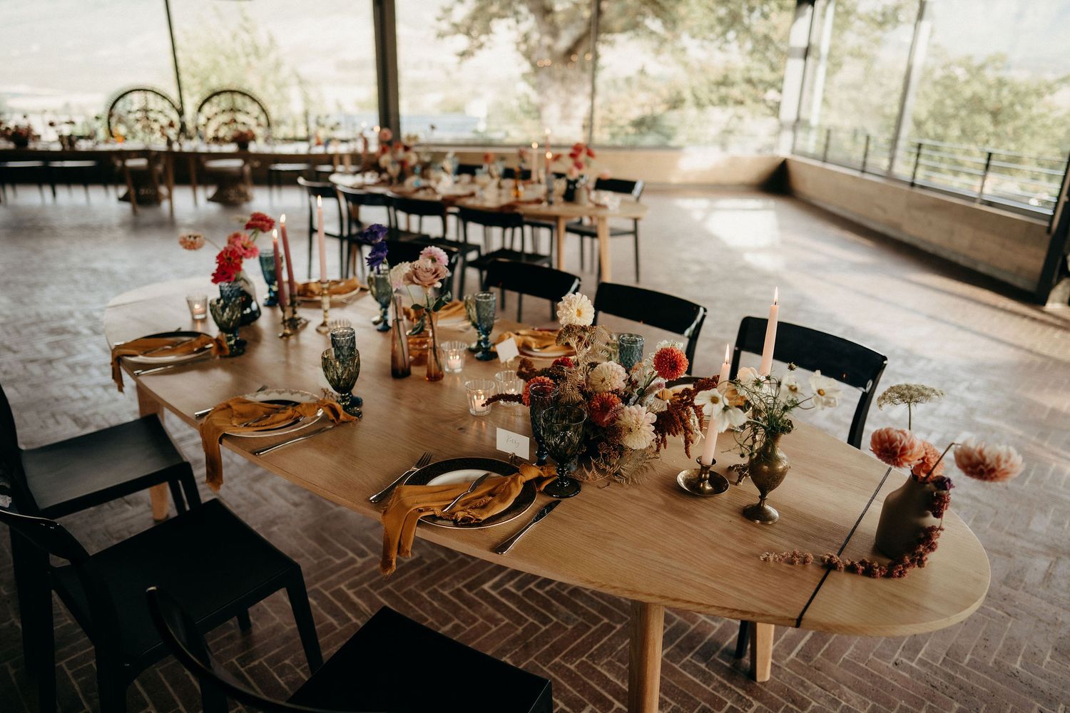 Rustic wooden dining table decorated with floral centerpieces and natural decor in an indoor-outdoor restaurant setting.