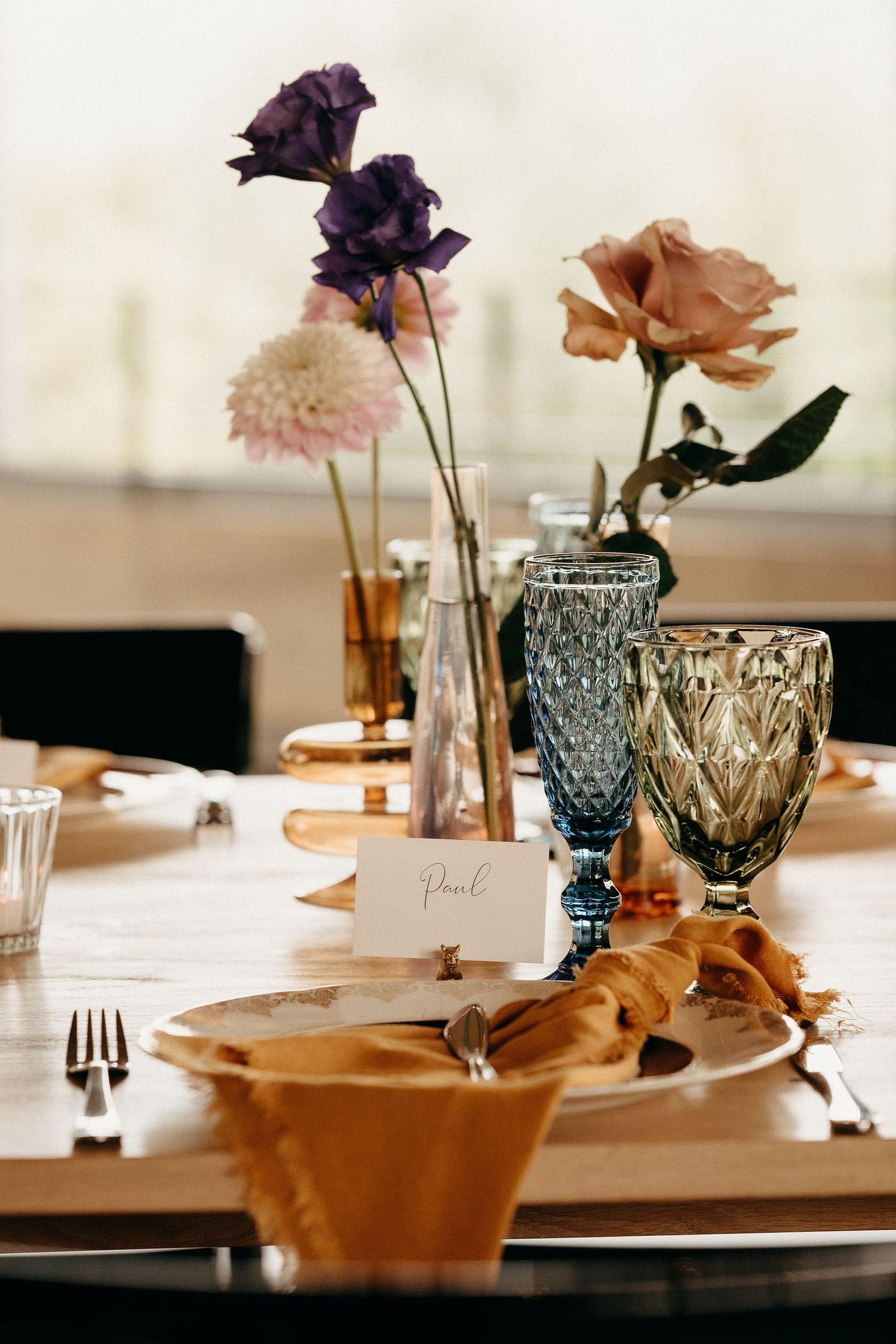 Elegant table setting with purple and peach flowers in textured glass vases alongside a place card and fabric napkin.